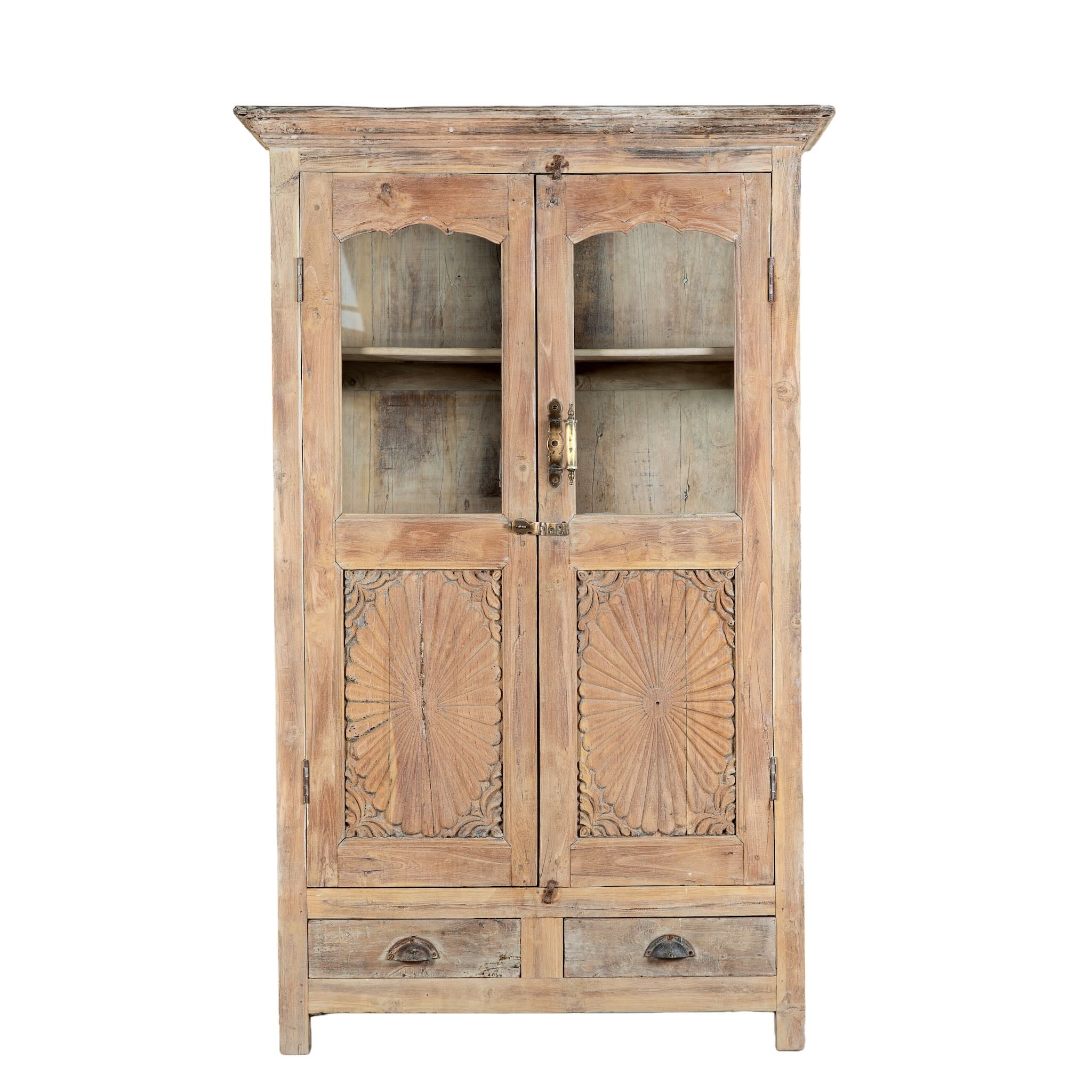 White background image of a reclaimed teak wood showcase Aasta with Mediterranean design, featuring hand-carved doors, glass panels, and drawers. A unique piece of refurbished Indian furniture offering elegant display and practical storage.