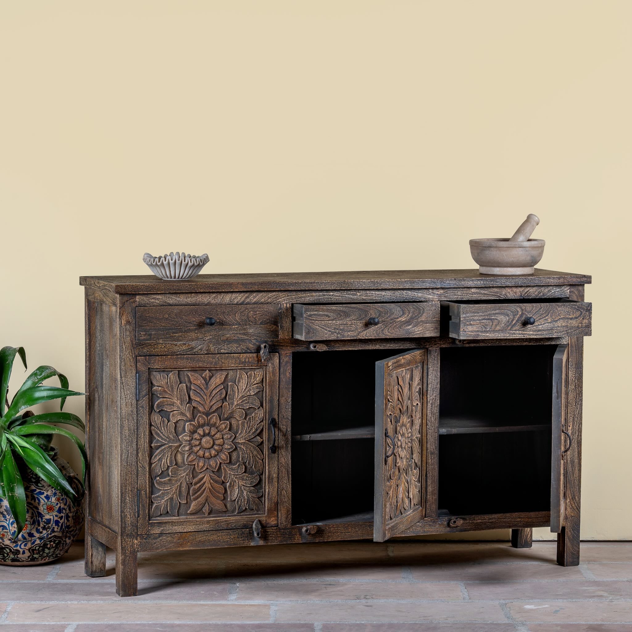 Side angle image with open doors of a dark brown mango wood sideboard with hand carved floral design, 3 drawers and 3 doors.