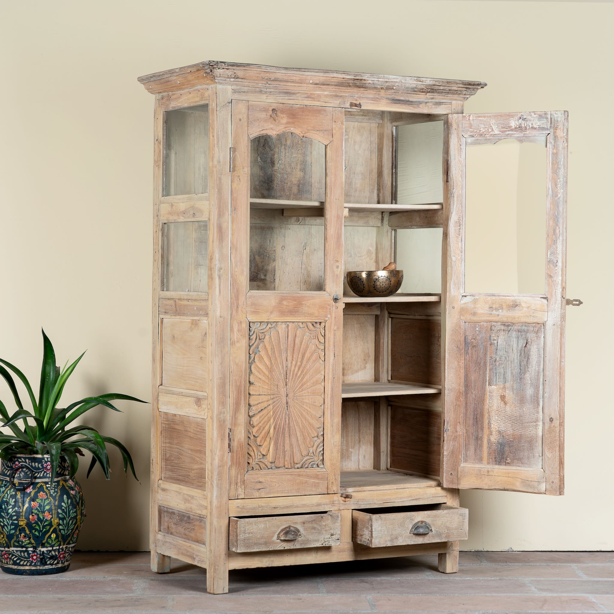 Side angle image with open doors of a reclaimed teak wood showcase Aasta with Mediterranean design, featuring hand-carved doors, glass panels, and drawers. A unique piece of refurbished Indian furniture offering elegant display and practical storage.