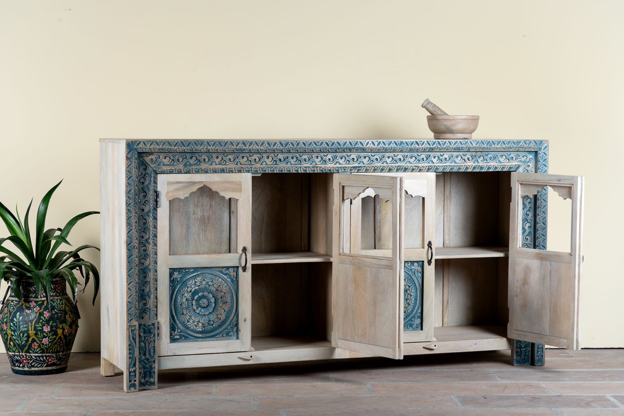 Side angle image with open doors of a hand carved showcase made of natural mango wood in white washed with blue floral accents and half glass doors in vintage design.