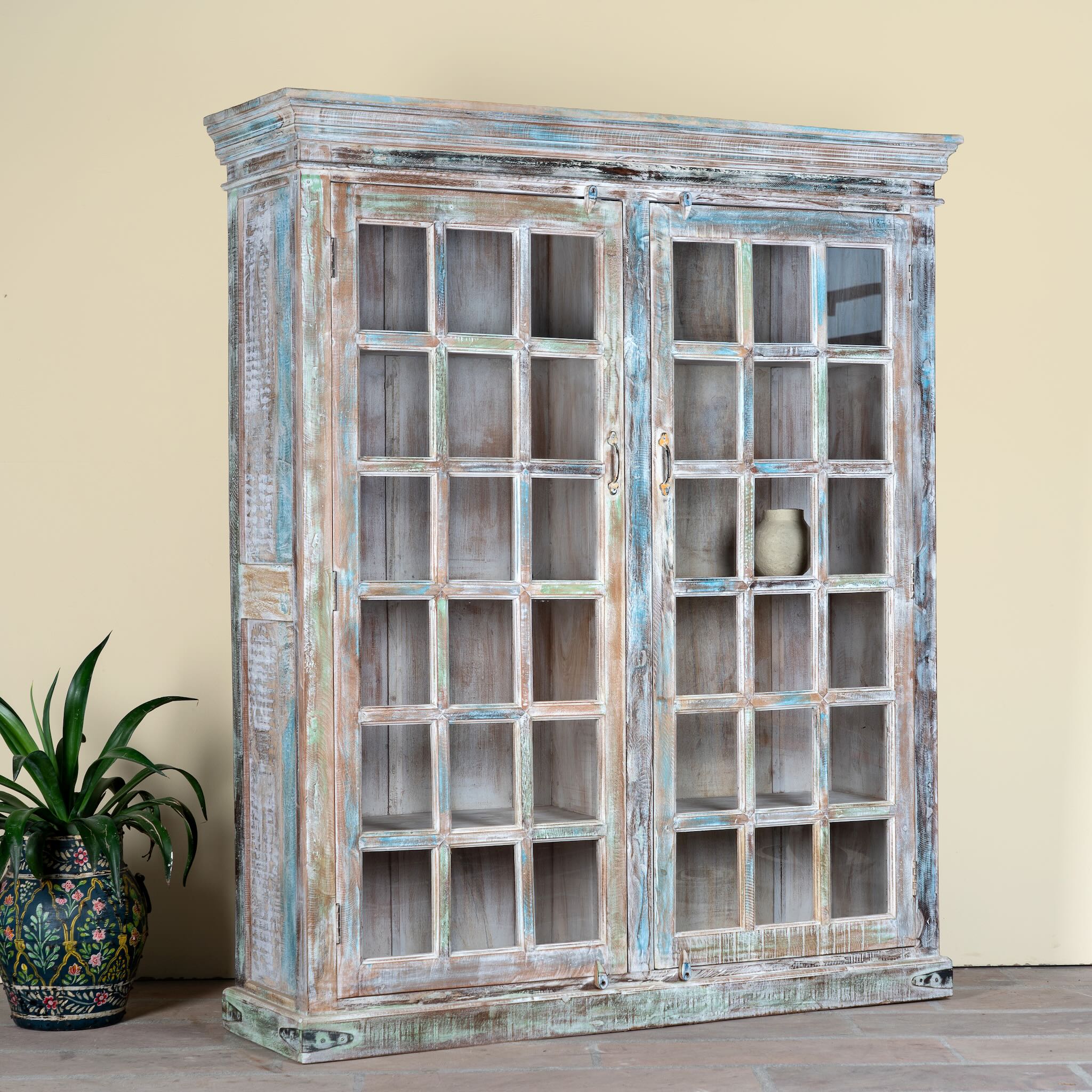 Side angle image of a reclaimed wood display cabinet Amara with distressed turquoise finish and glass-panelled doors. Sustainable living room furniture and vintage furniture piece with spacious shelving and authentic patina.