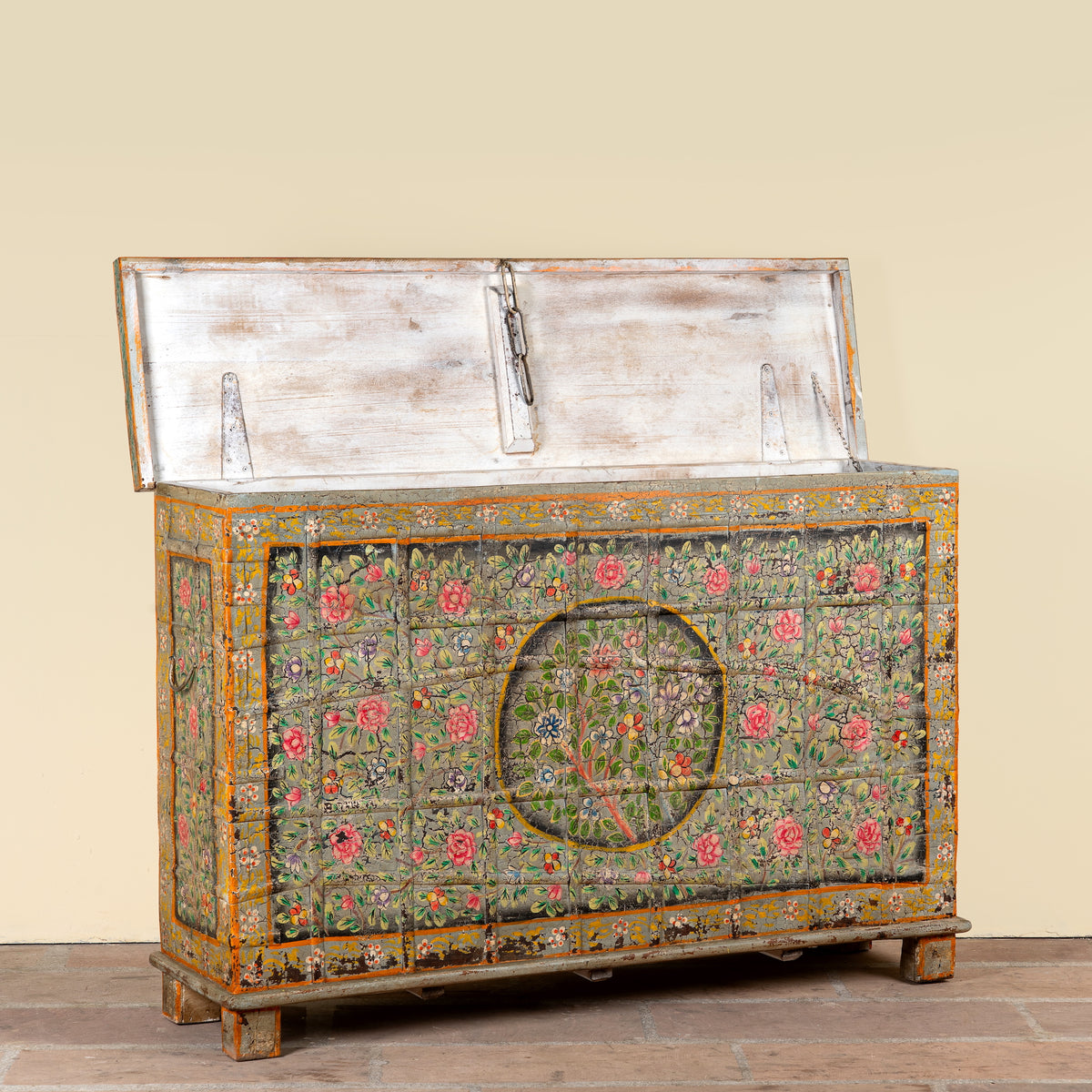 A distressed vintage wooden chest with a hinged lid, featuring hand-painted floral patterns in pink, red, and green on a weathered wood base, set against a plain beige wall.
