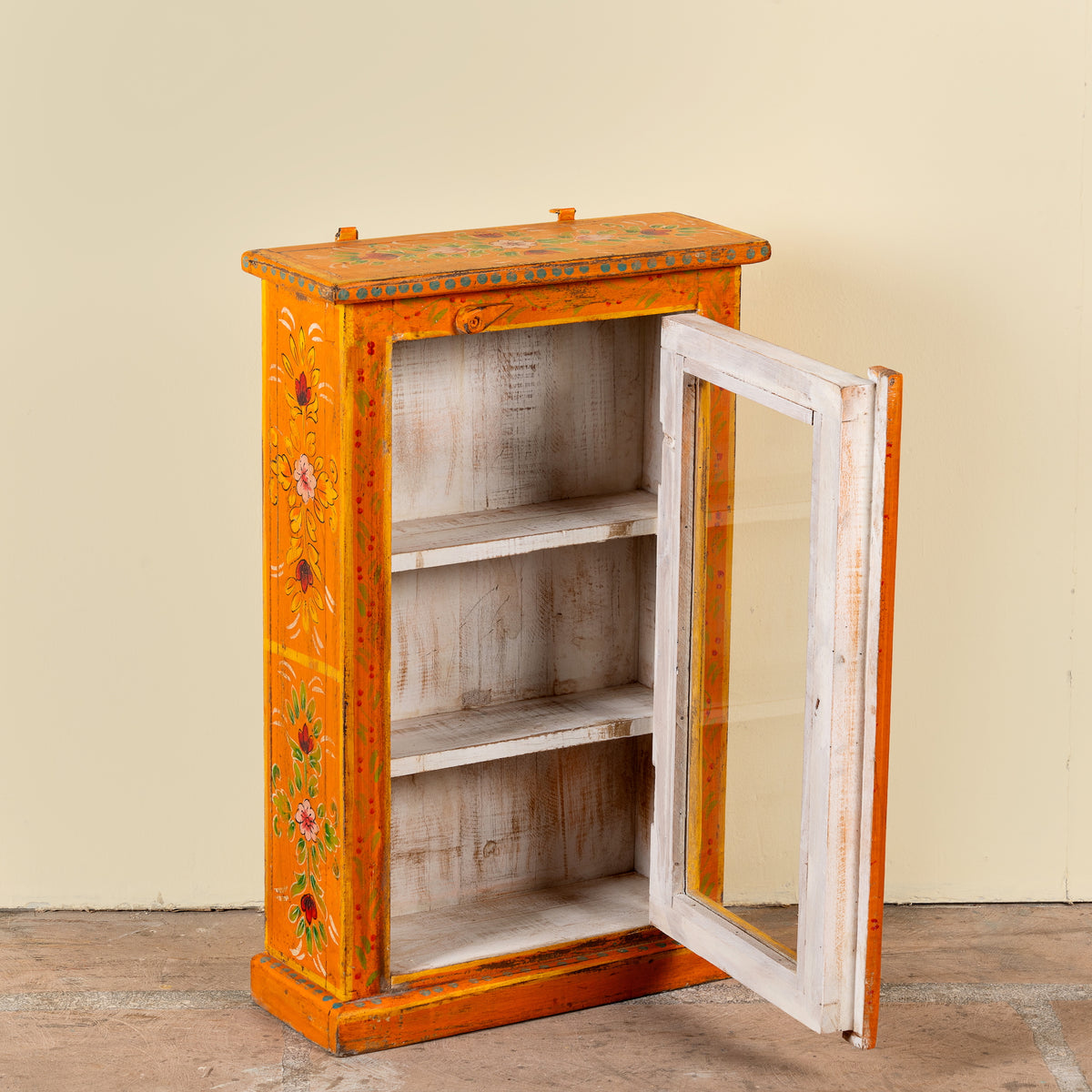 A small hand-painted orange cabinet with floral details, featuring a white distressed wooden frame door with a glass panel, three interior shelves, and a rustic finish, set against a plain beige wall.