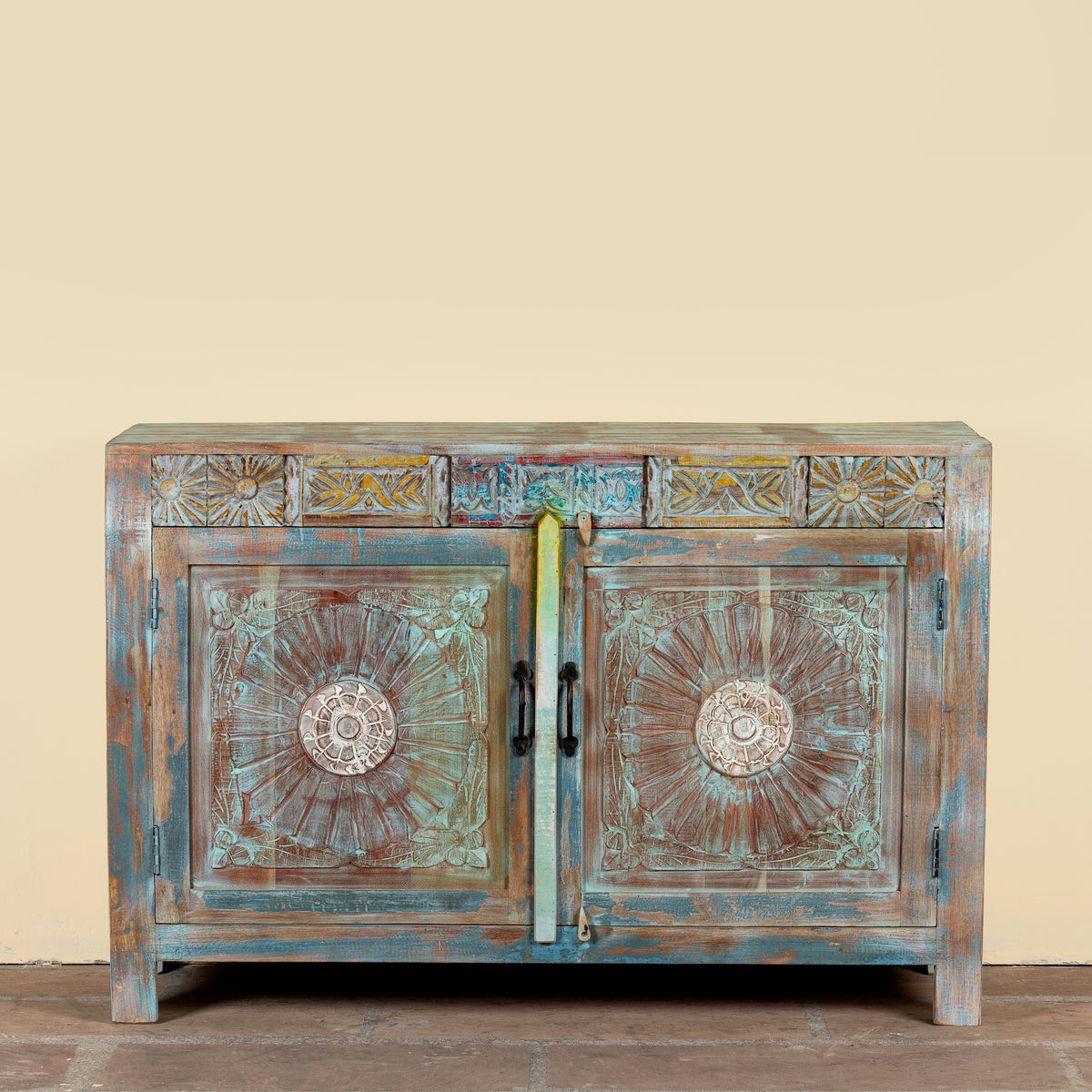 A rustic, hand-painted wooden sideboard cabinet with distressed teal, brown, and gold tones. The cabinet features two doors with ornate circular medallion carvings, decorative panels above, and a distressed finish.