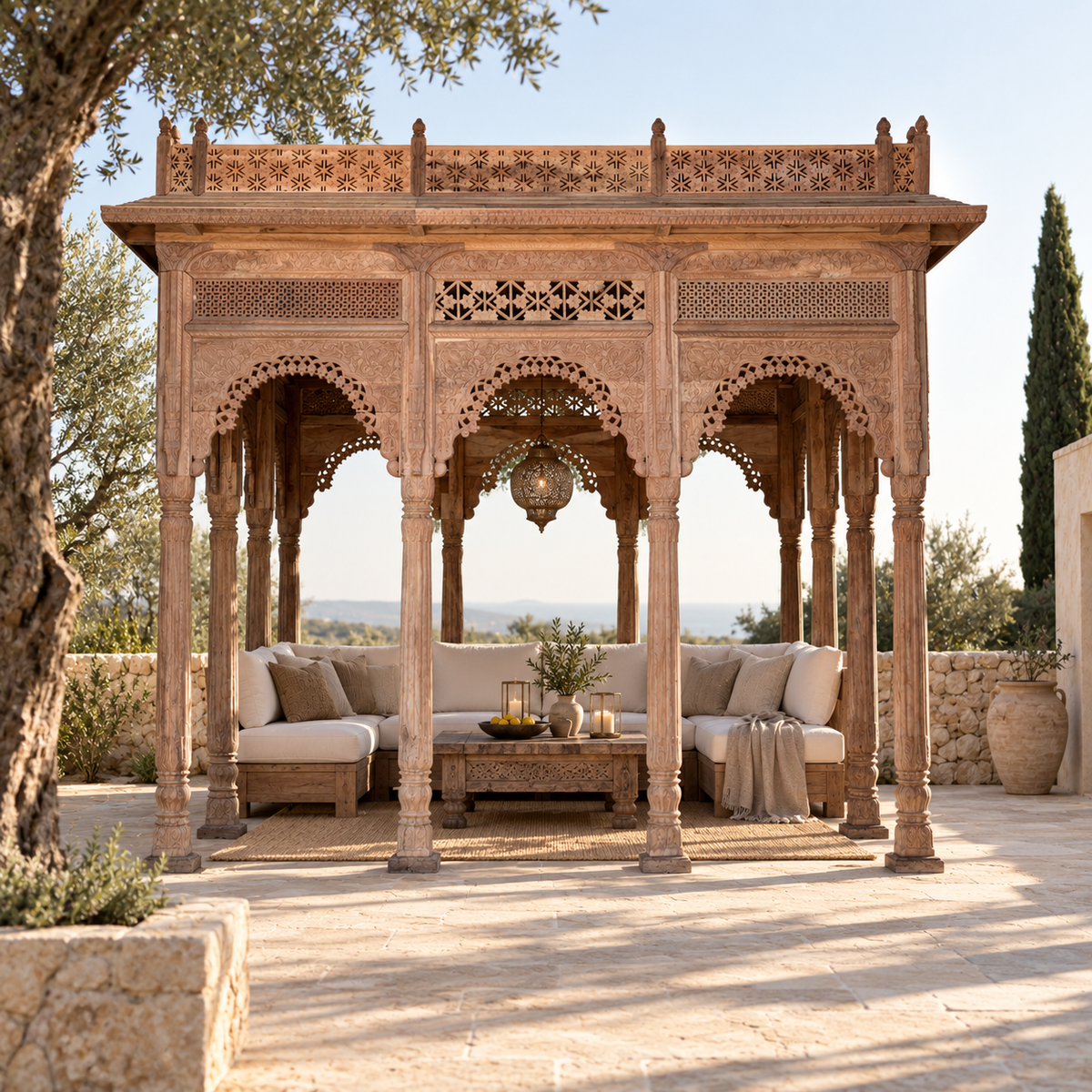 An ornate carved wooden outdoor pavilion with multiple arched openings and columns, furnished with a light beige sectional sofa, matching cushions, and a low wooden coffee table on a sunlit patio.