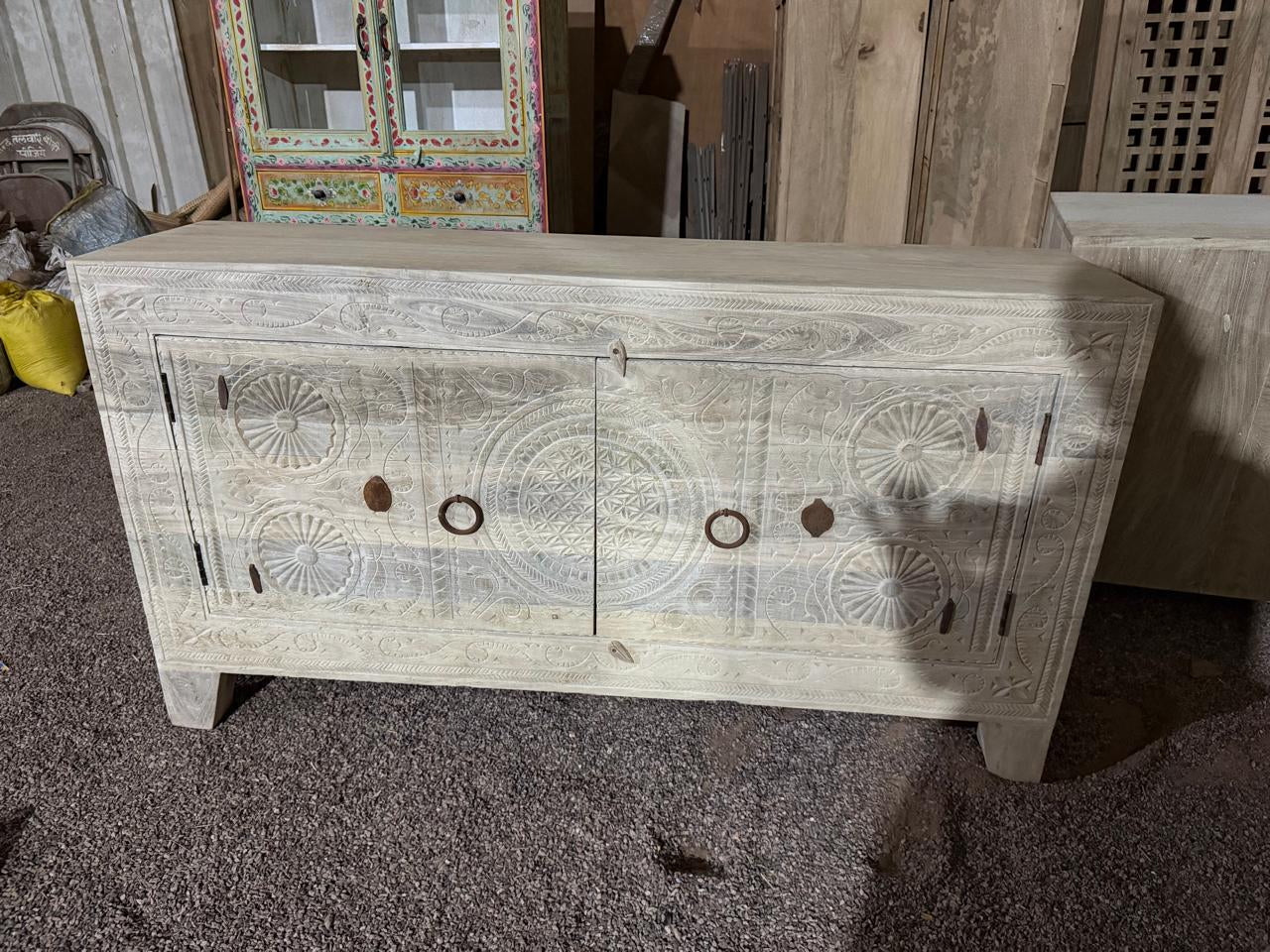 A carved wooden sideboard cabinet in a weathered off-white finish, featuring circular and floral engravings and rustic metal ring pulls, photographed in a workshop or warehouse setting.