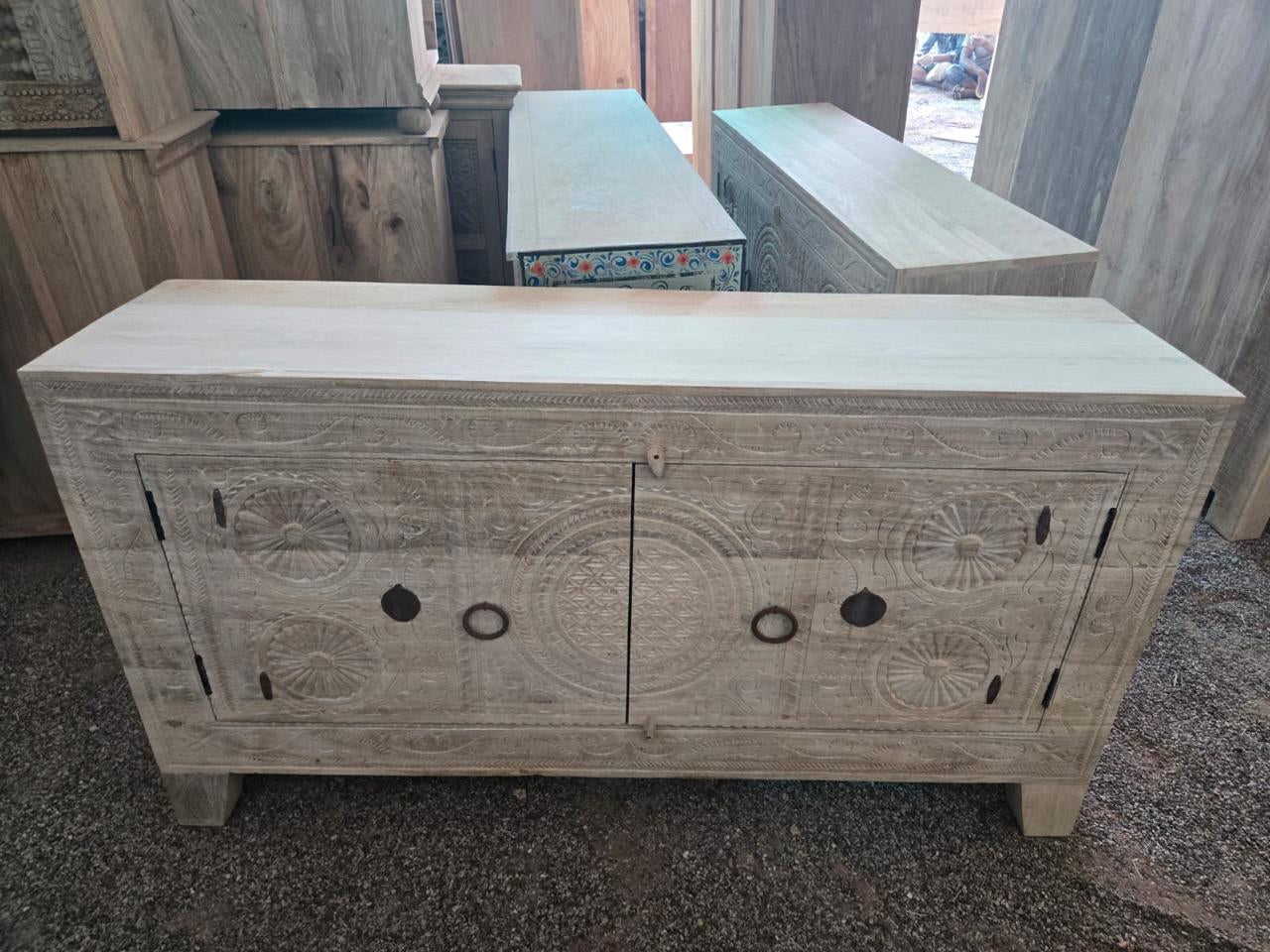 A carved light beige wooden sideboard cabinet with circular patterns on the doors and dark metal ring pulls, photographed in a showroom with other furniture pieces in the background.