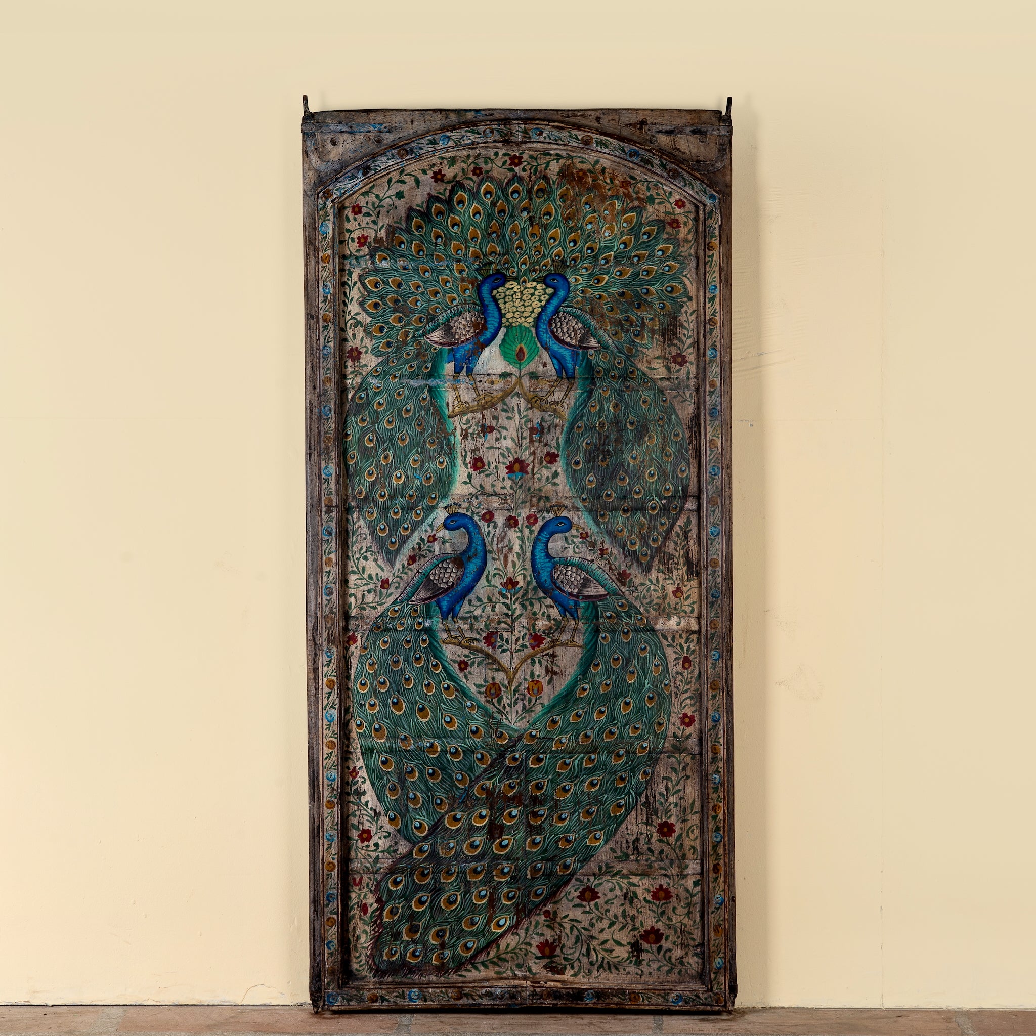 A tall, narrow hand-painted wooden door panel featuring two peacocks with elaborate teal, blue, and gold feather patterns on a distressed brown background, set against a plain beige wall.