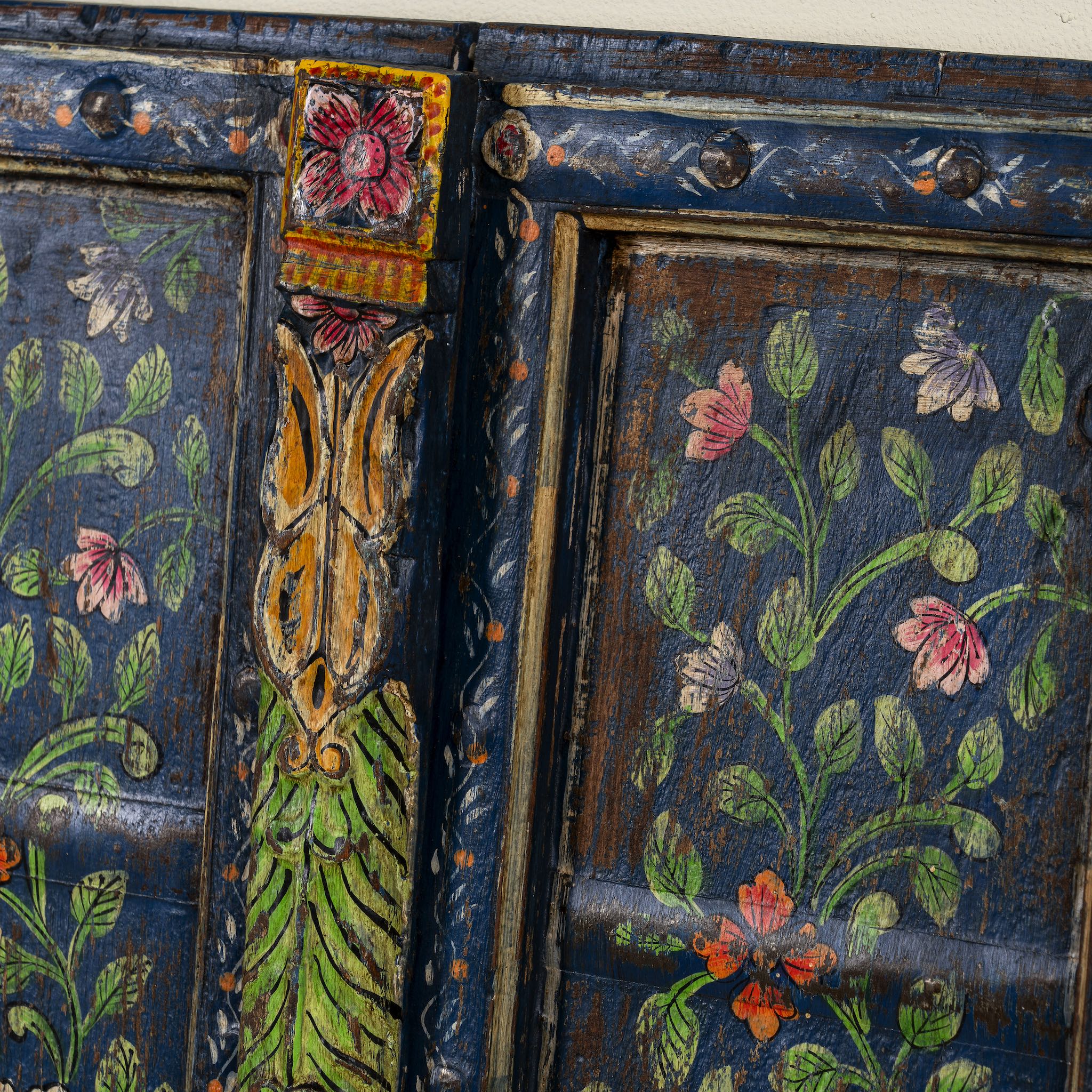 Close up image of a hand-painted wall panel made from an old teak wood window frame, adorned with detailed floral motifs in shades of pink, red, green, and white against a deep blue background, framed by intricate carvings and colourful decorative accents.