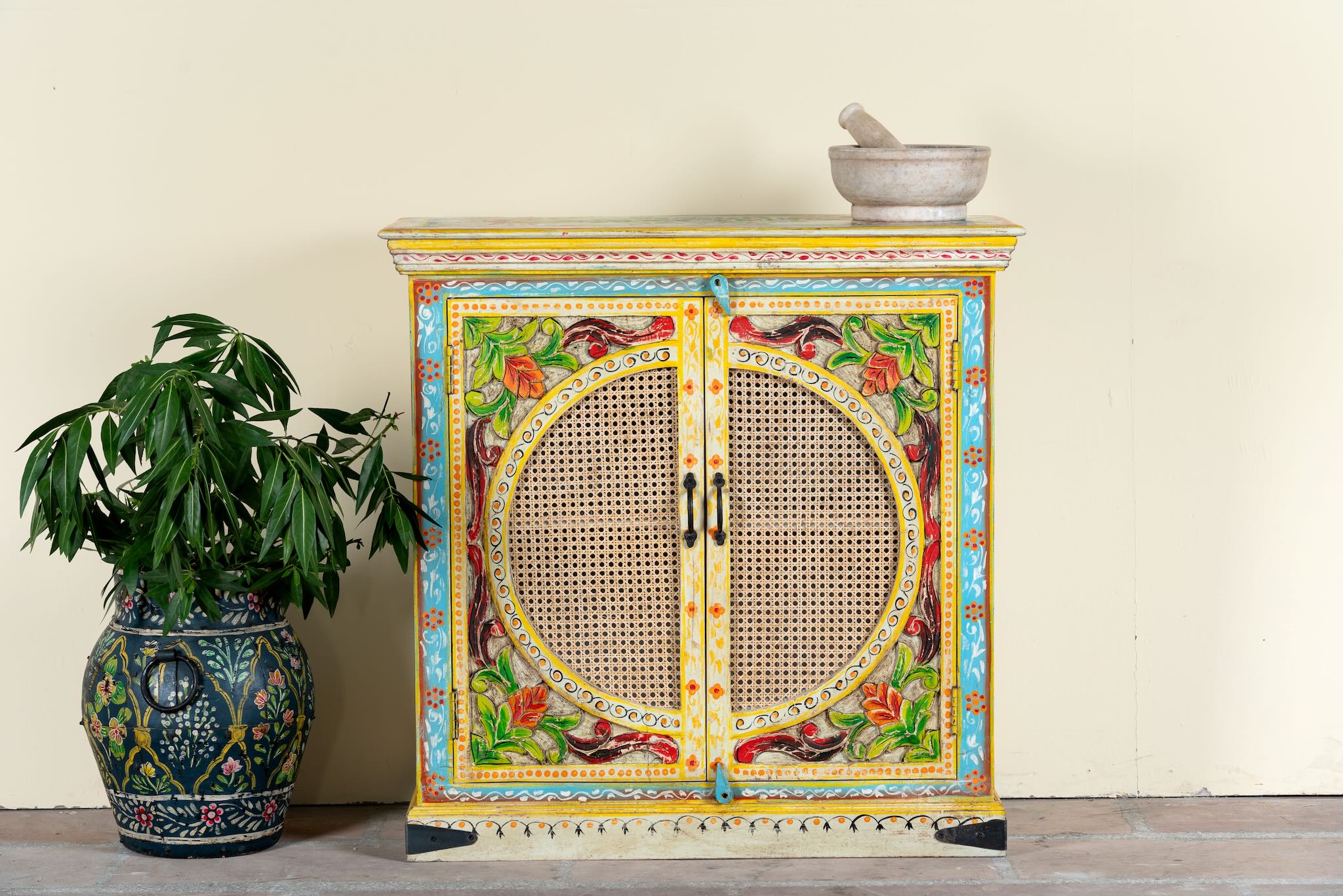 White background image of a hand-painted, hand-carved cupboard made of mango wood with floral design and rattan inserts.
