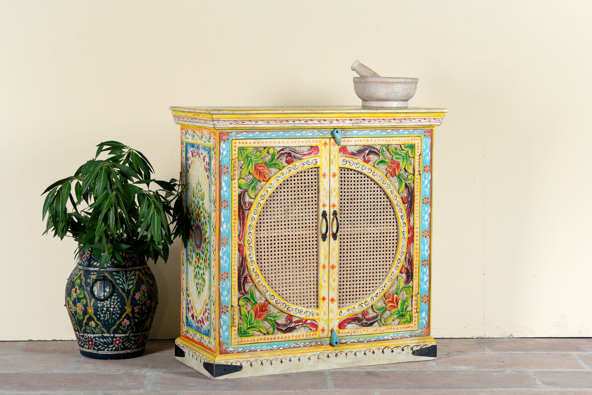 Side angle image of a hand-painted, hand-carved cupboard made of mango wood with floral design and rattan inserts.