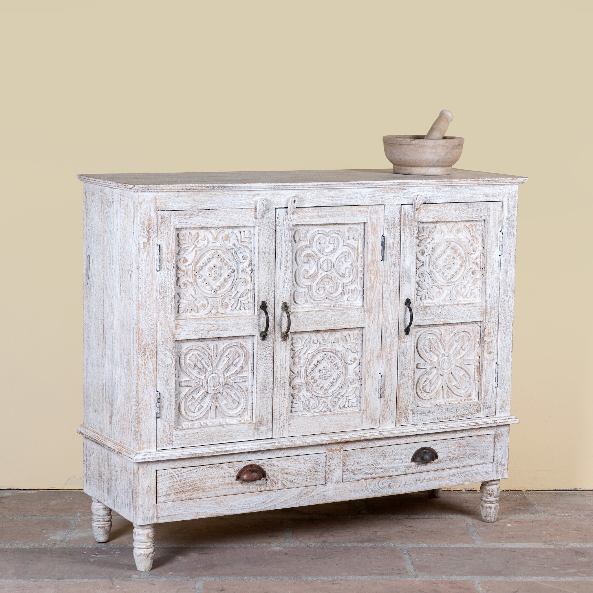 Side angle image of a whitewashed hand carved wooden sideboard made from solid mango wood with floral patterns, 3 doors and 2 drawers; artisanal mango wood furniture, 120 × 100 × 40 cm.