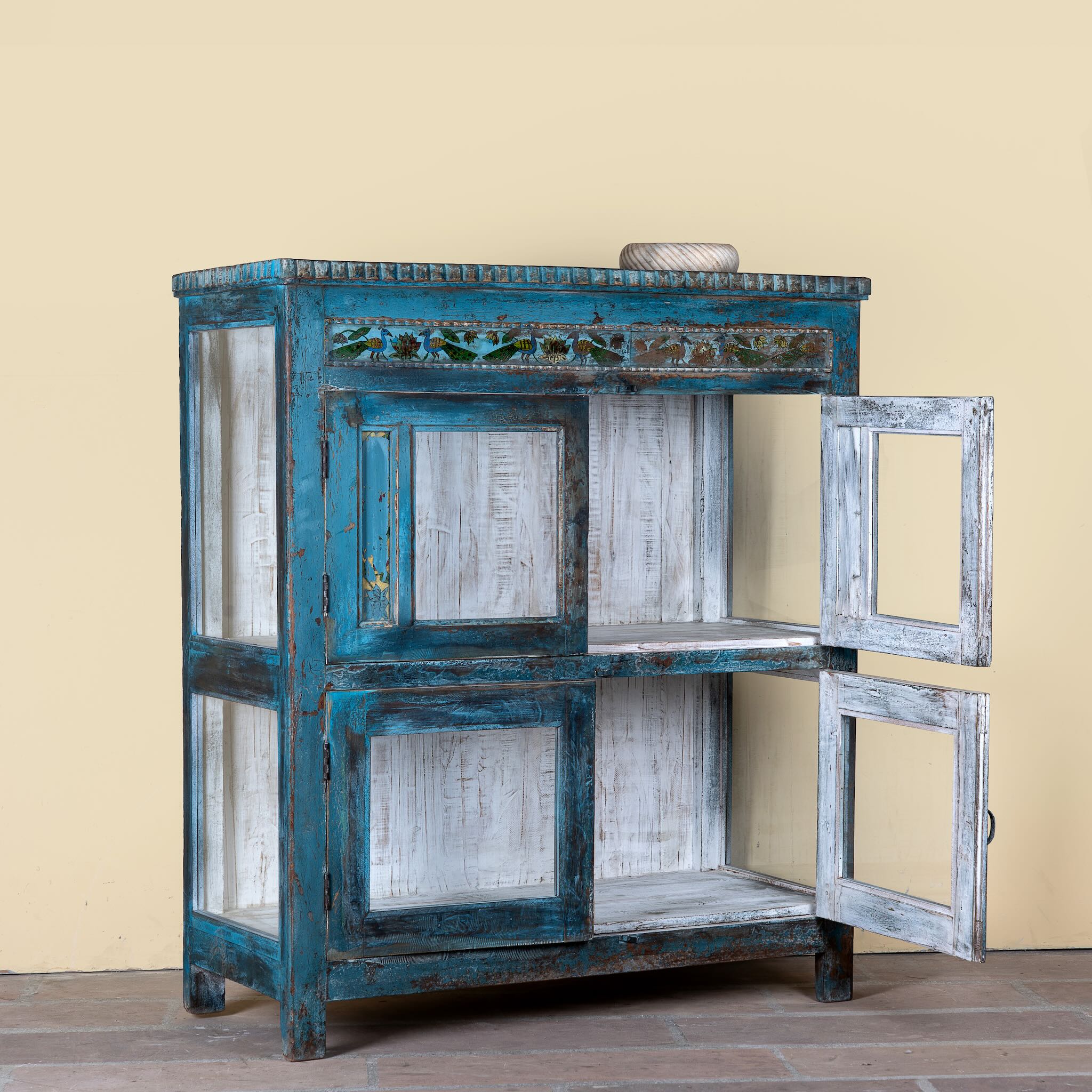 Side angle image with open door of a Kavish vintage blue reclaimed teak wood wardrobe with peacock reverse glass paintings and distressed Indian finish.