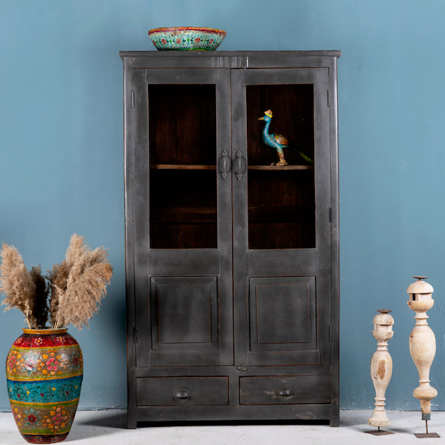 A shabby chic-style reclaimed teak wood showcase cabinet with glass doors and various decorative items displayed on its shelves, set against a plain blue wall.