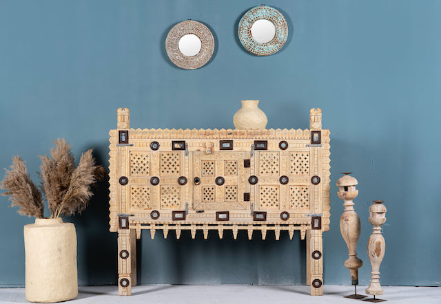 An antique Indian treasure chest made of solid teak wood with a sandblasted finish, featuring hand-carved patterns and iron applications, displayed with decorative items and mirrors in the background.