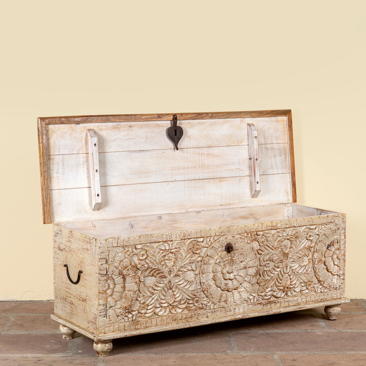 A rustic carved wooden storage chest with a distressed cream and brown finish, featuring floral carvings on the front, a hinged lid, and metal hardware, set against a solid beige wall.