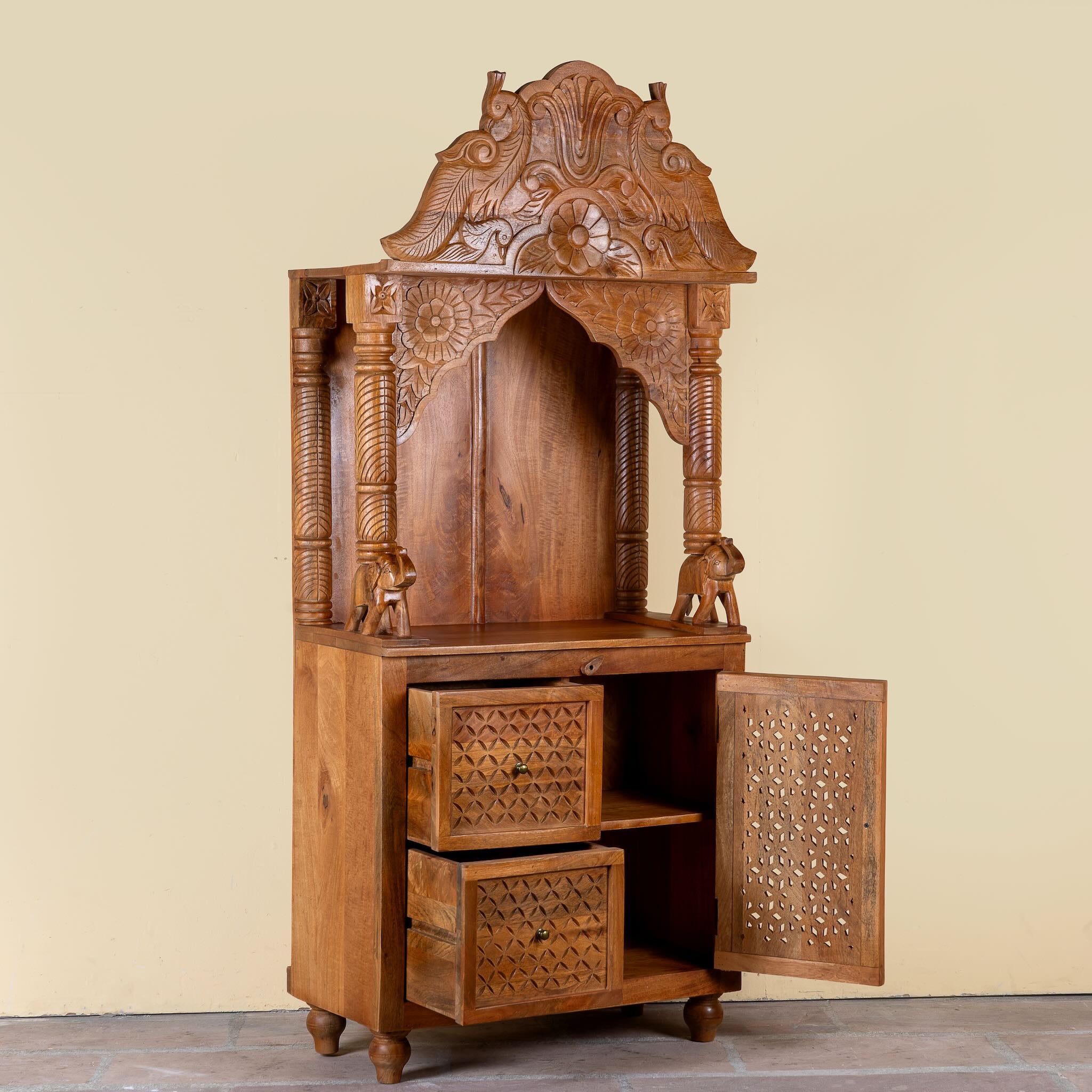 Side angle image with open door of a Moksha hand carved mango wood mandir with decorative Indian carvings, drawers, and cabinet storage.