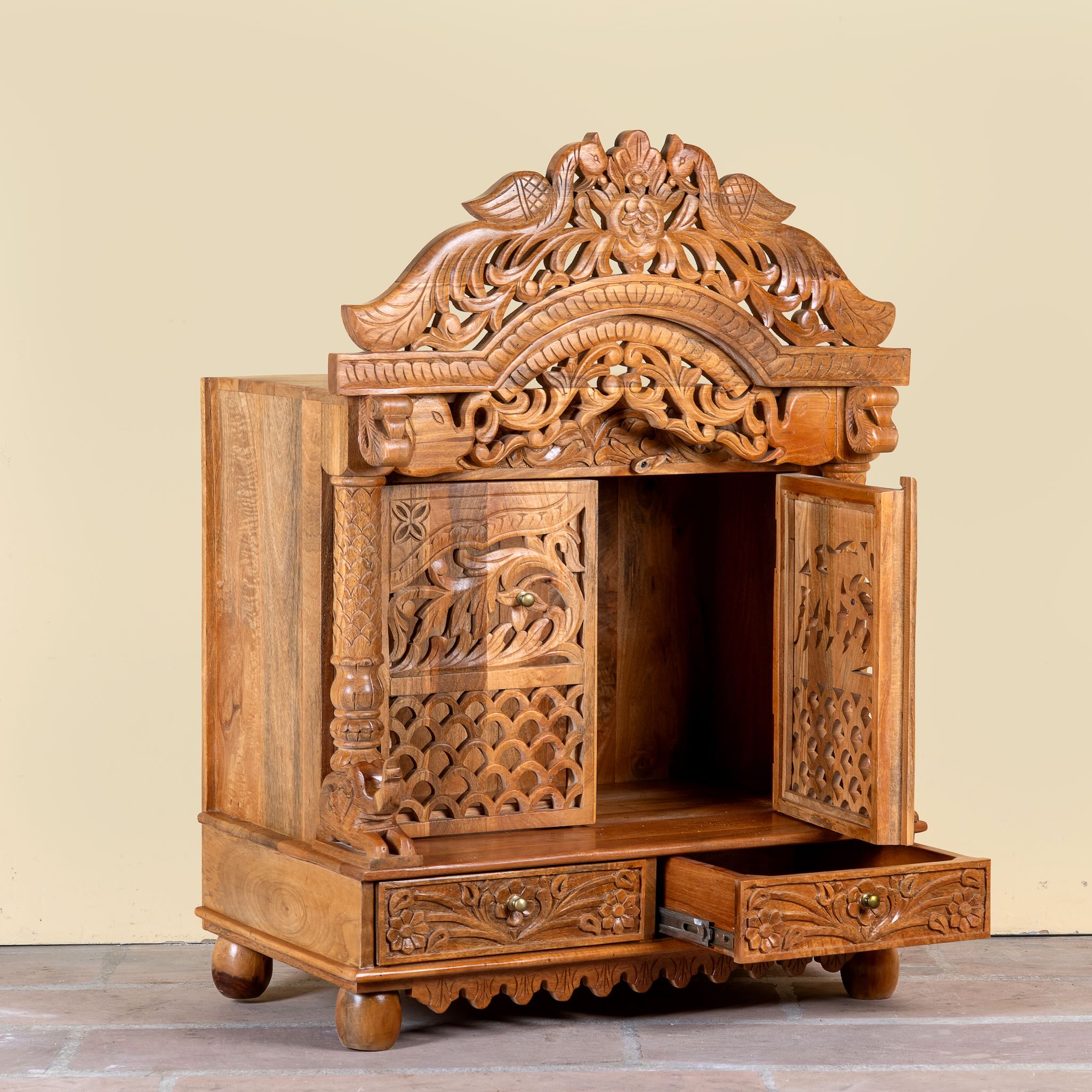 Side angle image of a hand carved mango wood Indian mandir temple with ornate floral carvings, lattice doors, and two storage drawers.