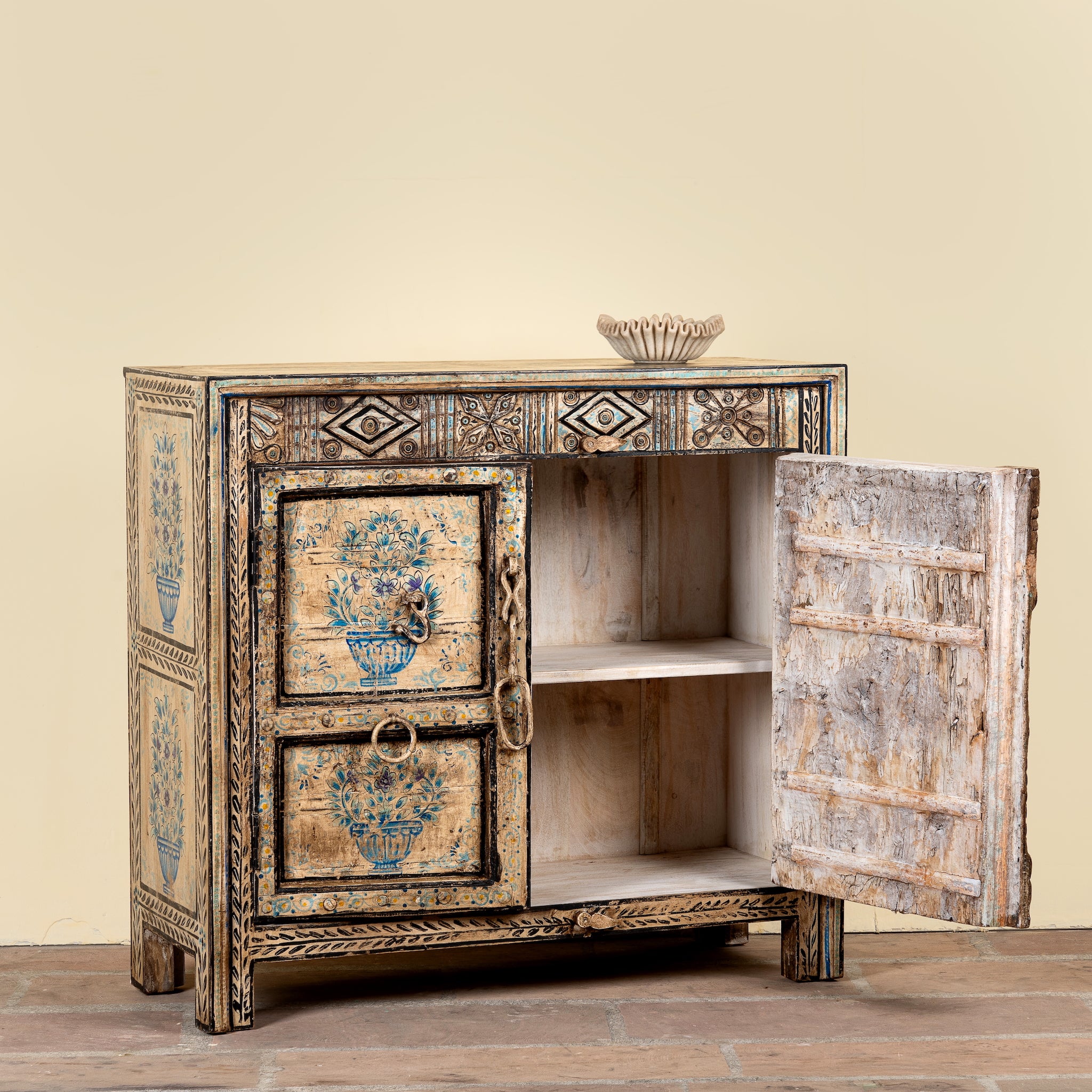 A rustic hand-painted wooden cabinet with a distressed finish, featuring blue floral motifs on the doors and ornate geometric carvings. The cabinet has one open door showing an interior shelf, and a small decorative bowl sits on top against a solid beige background.