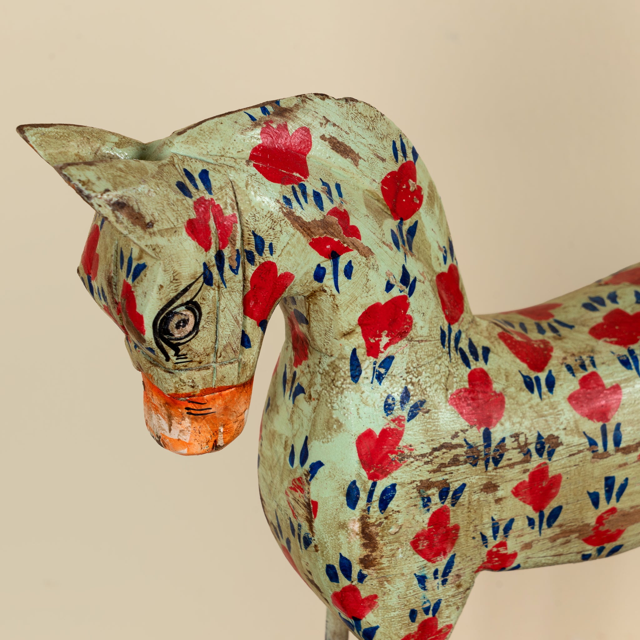A decorative wooden horse figurine with a distressed light green finish, decorated with hand-painted red flowers, blue leaves, and an orange muzzle, shown against a solid beige background.