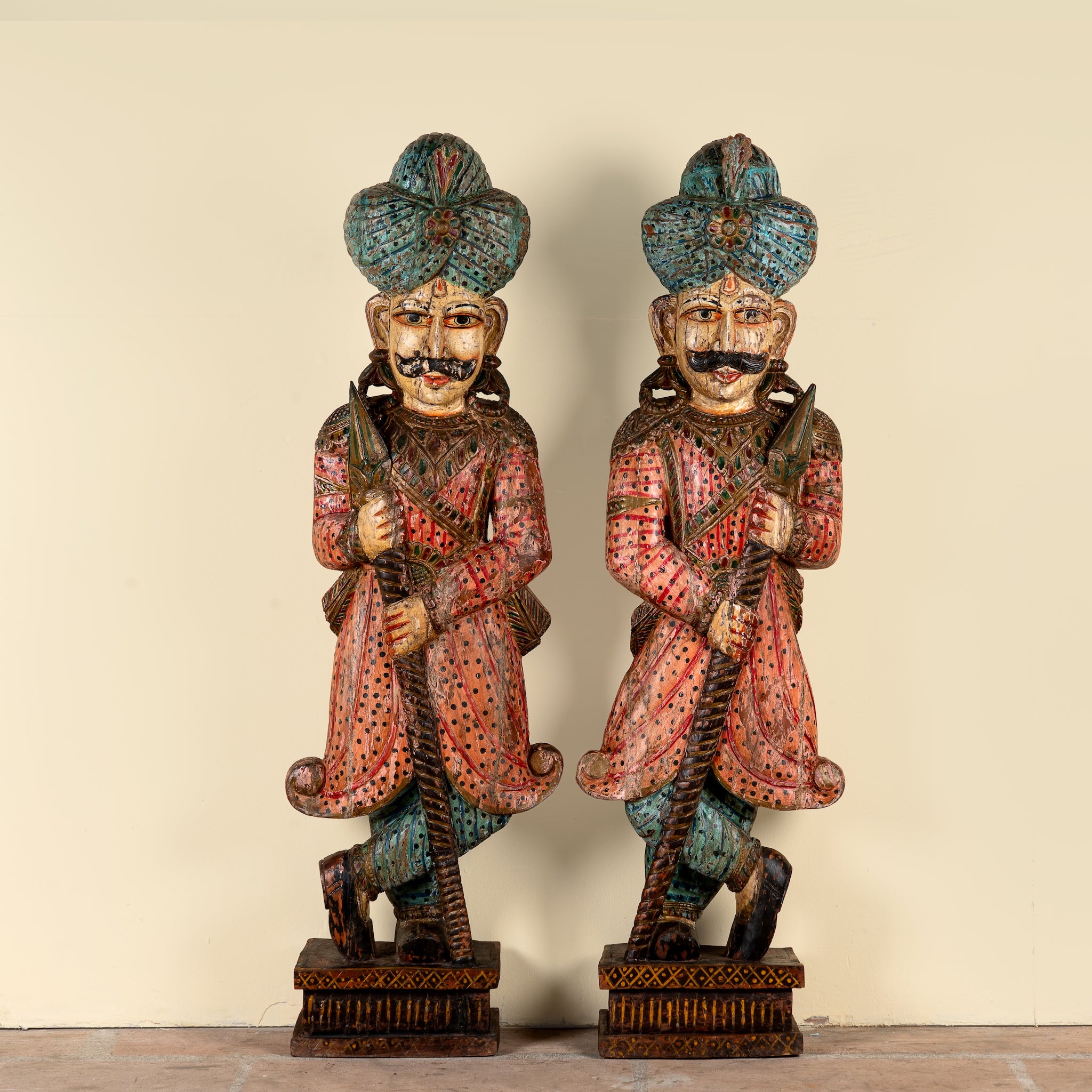 A pair of hand-carved wooden Indian warrior statues with turquoise, red, and black painted details, standing on a beige background.