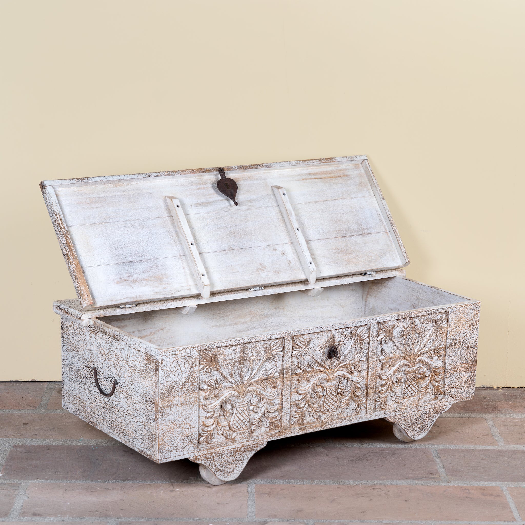 Side angle with open lid of a Shaan mango wood storage chest coffee table with carved front, iron fittings, wooden wheels and cream distressed finish 116cm