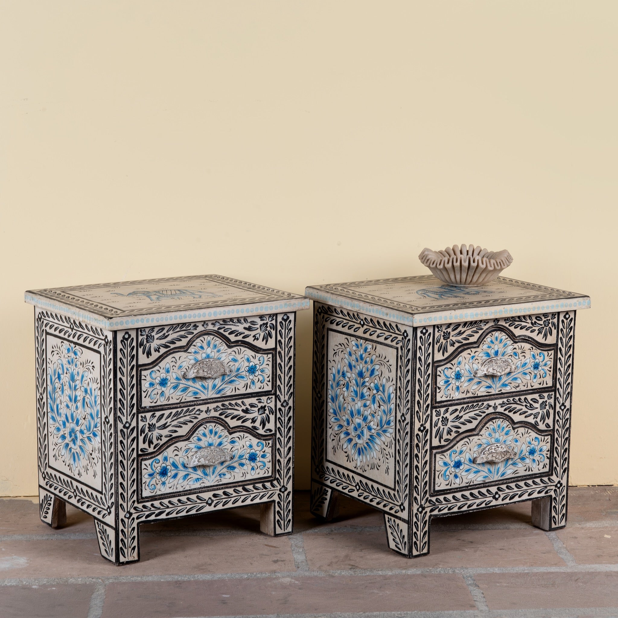 Side angle image of a pair of hand-painted nightstands made from reclaimed wood, featuring floral motifs in soft turquoise and black with intricate carved detailing and two drawers for storage.