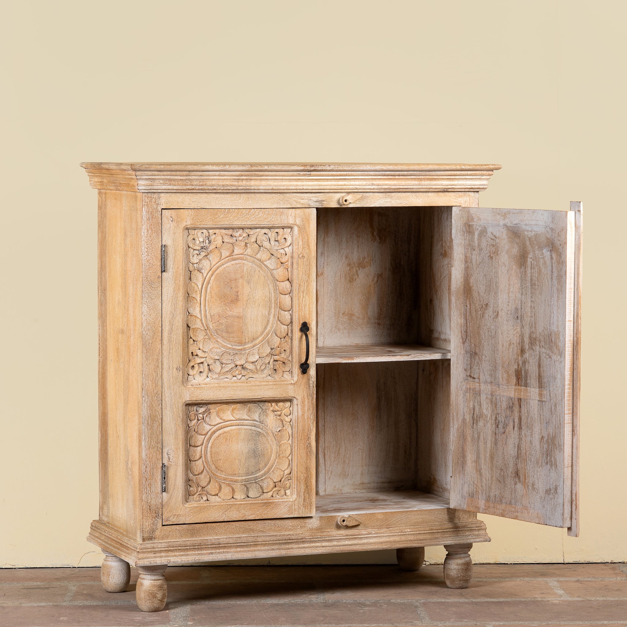 A small carved wooden cabinet with one door open, showing an interior shelf. The cabinet has ornate floral and circular carvings on the left door and a distressed, weathered finish.