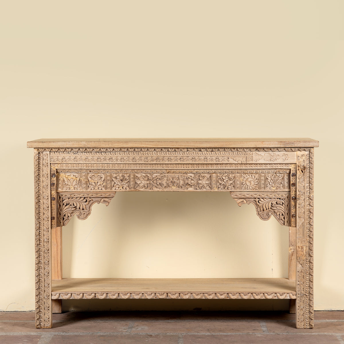An ornate carved wooden console table with a natural light wood finish, featuring intricate floral carvings on the apron, legs, and lower shelf, set against a plain beige wall.