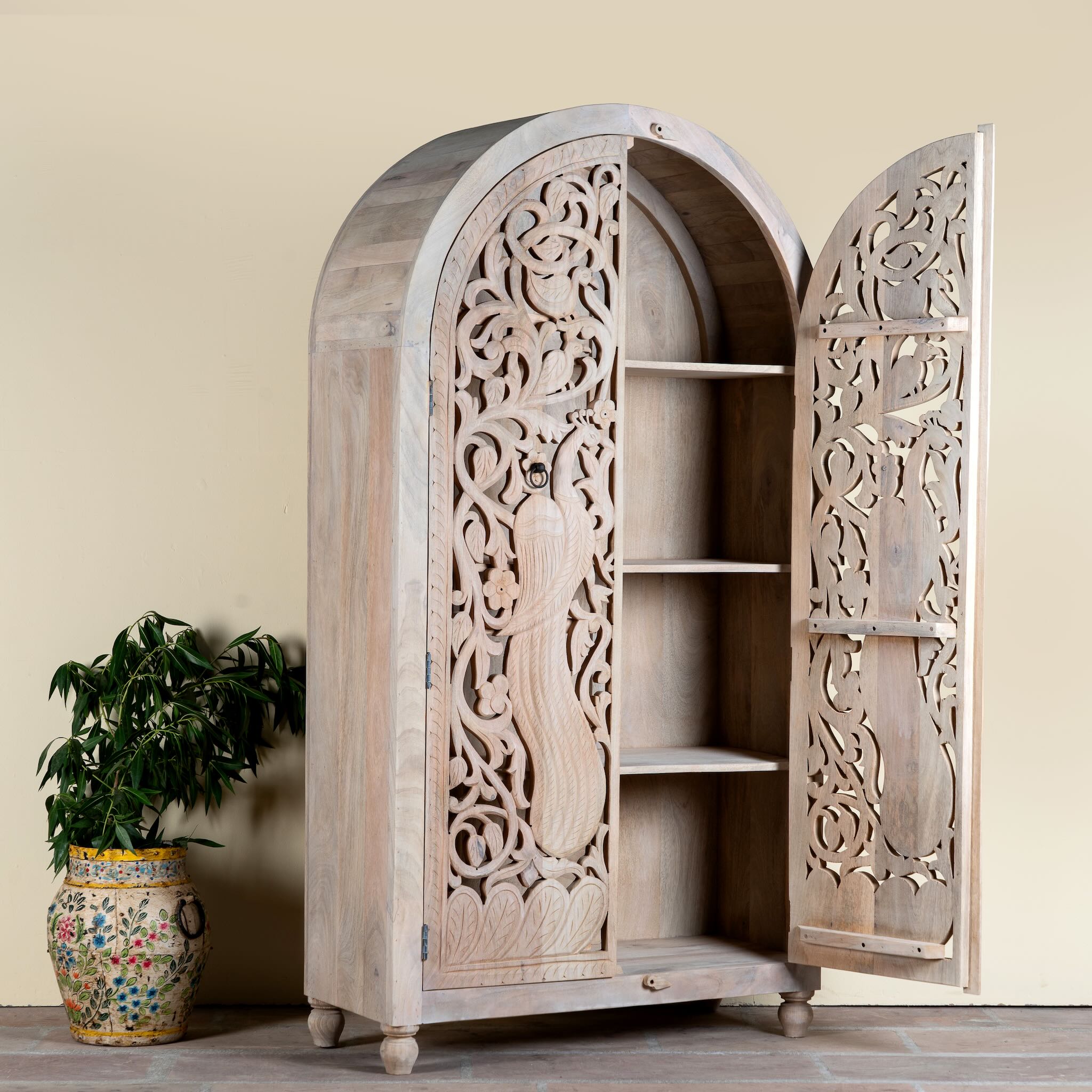 Side angle image with open door of a hand-carved mango wood wardrobe with arched top and intricate peacock floral design – artisanal Indian bedroom furniture.