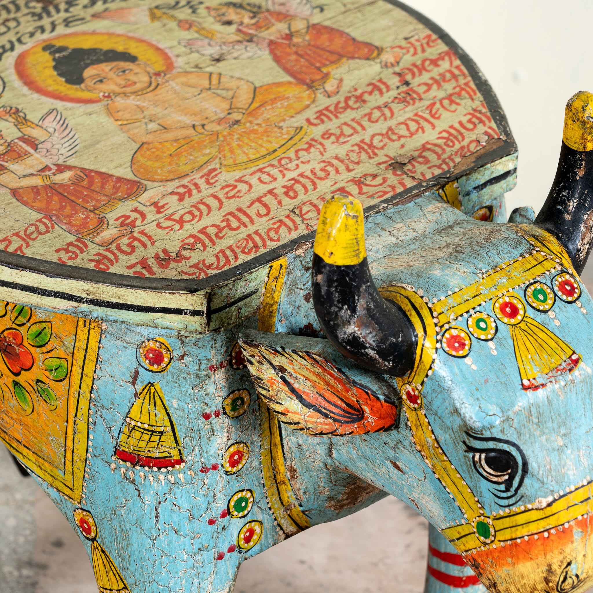 Close up image of a hand-painted wooden cow-shaped side table with Indian script and blue folk art patterns, perfect for vintage and India-inspired home interiors.