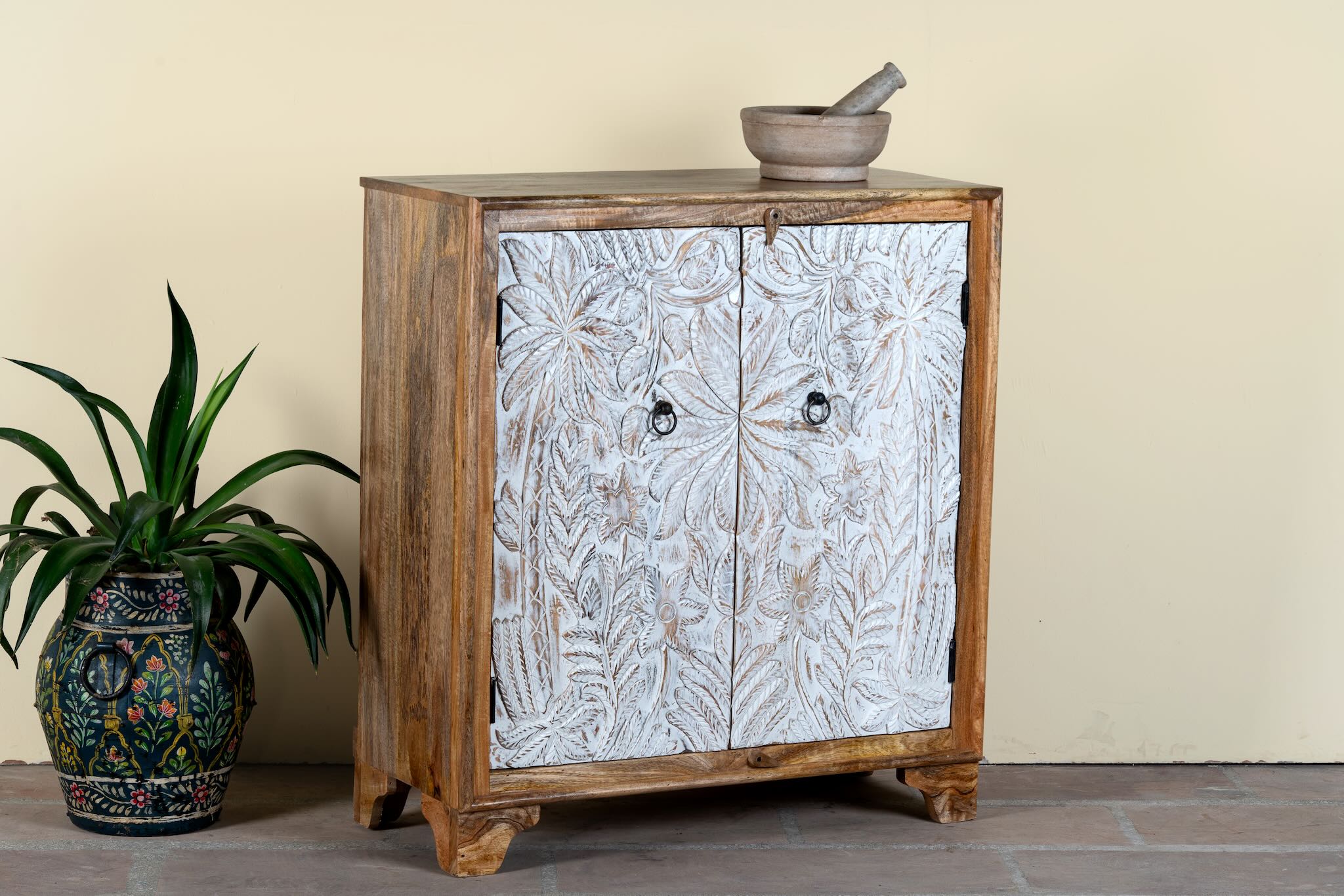 Side angle image of a hand-carved mango wood cupboard Utkarsh with whitewashed doors featuring palm tree and botanical motifs. A Mediterranean-inspired piece of Indian furniture combining artisan detail with practical storage.