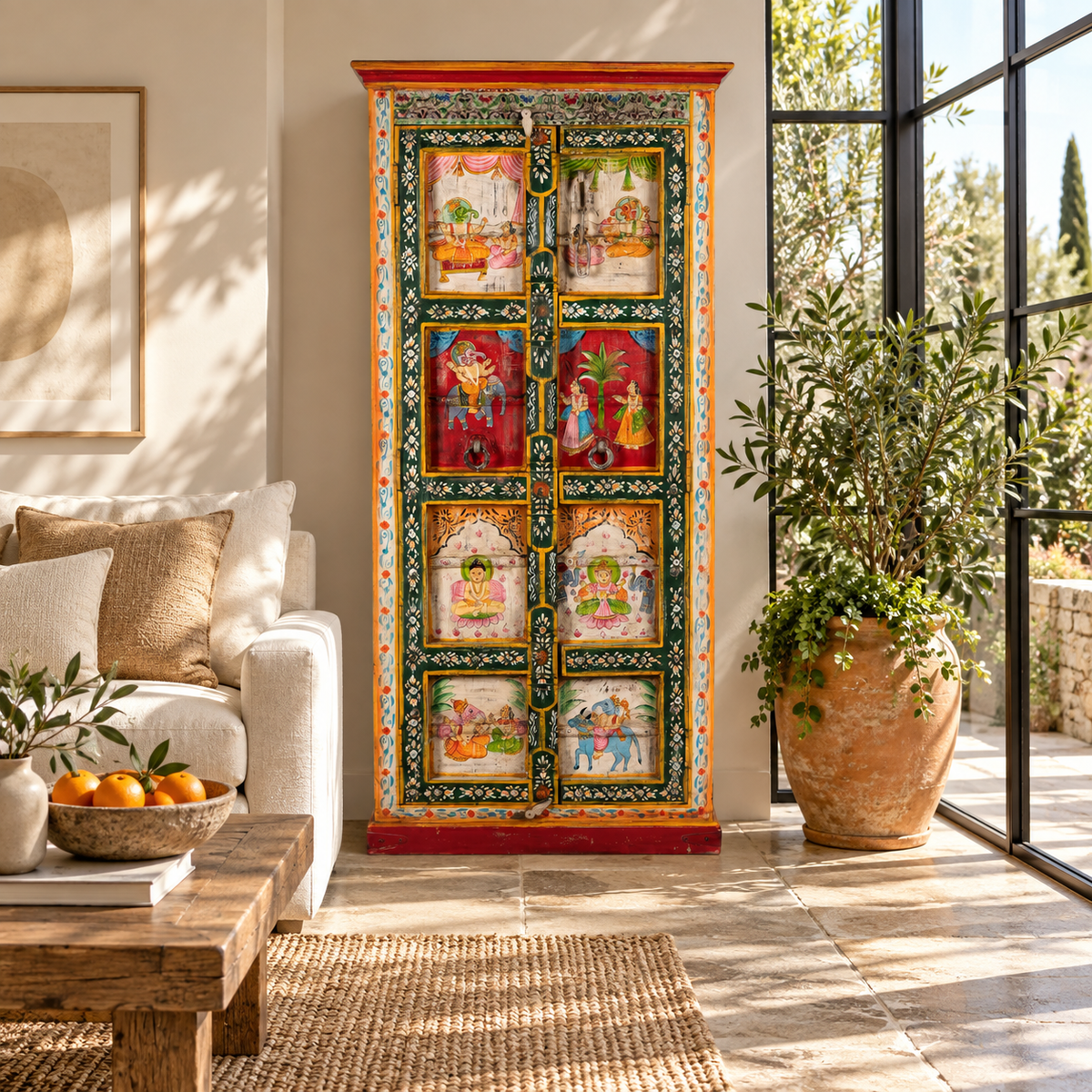 Lifestyle image of a tall, hand-painted Indian wooden cabinet with colorful traditional illustrations, set in a bright, modern living room next to a large potted plant.