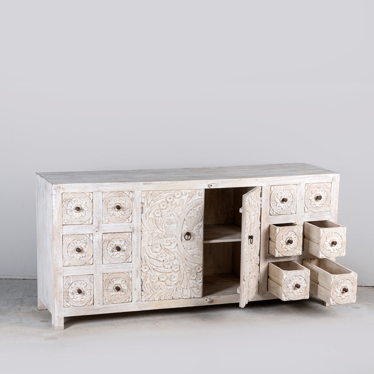 A carved wooden sideboard cabinet in a distressed off-white finish, featuring multiple small drawers on both ends, a central carved door, and one open door with additional drawers pulled out to the right, set against a plain gray studio background.