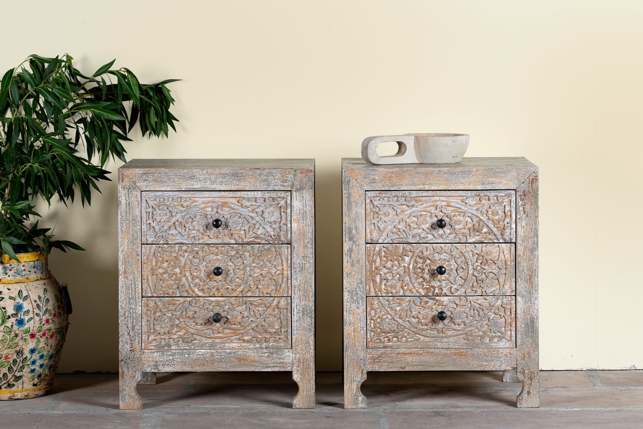 Lifestyle image of a pair of hand-carved mango wood nightstands Vikram with three drawers each, showcasing intricate floral motifs in a distressed vintage-style finish. Elegant and functional bedside tables with timeless artisan appeal.