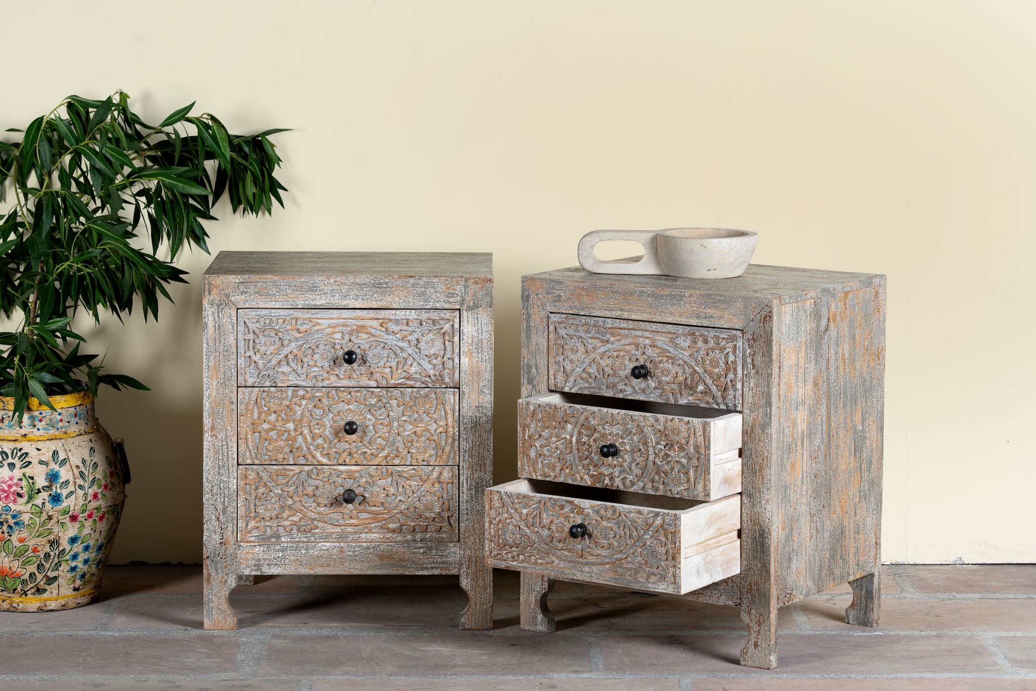 Side angle image of a pair of hand-carved mango wood nightstands Vikram with three drawers each, showcasing intricate floral motifs in a distressed vintage-style finish. Elegant and functional bedside tables with timeless artisan appeal.