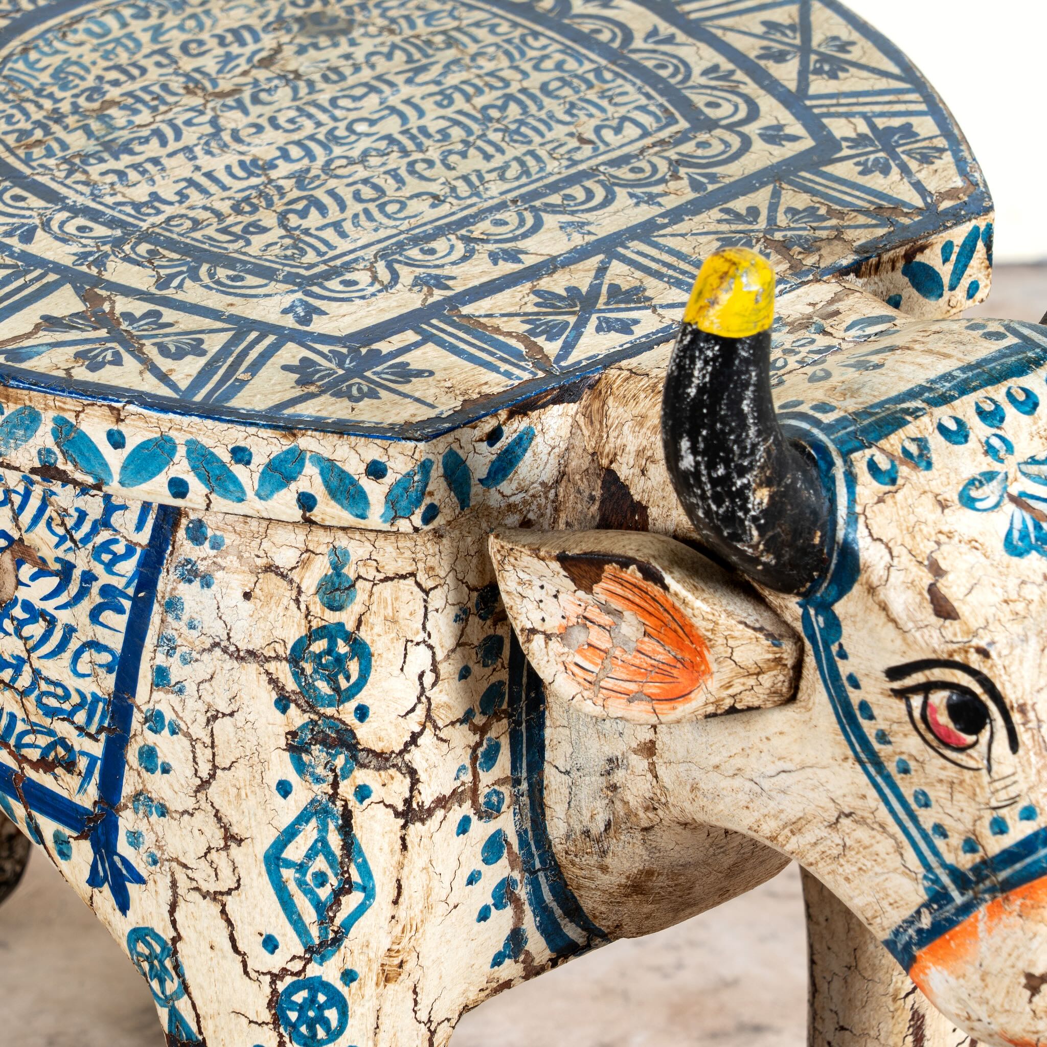 Close up image of a hand-painted wooden cow-shaped side table with Indian script and blue folk art patterns, perfect for vintage and India-inspired home interiors.