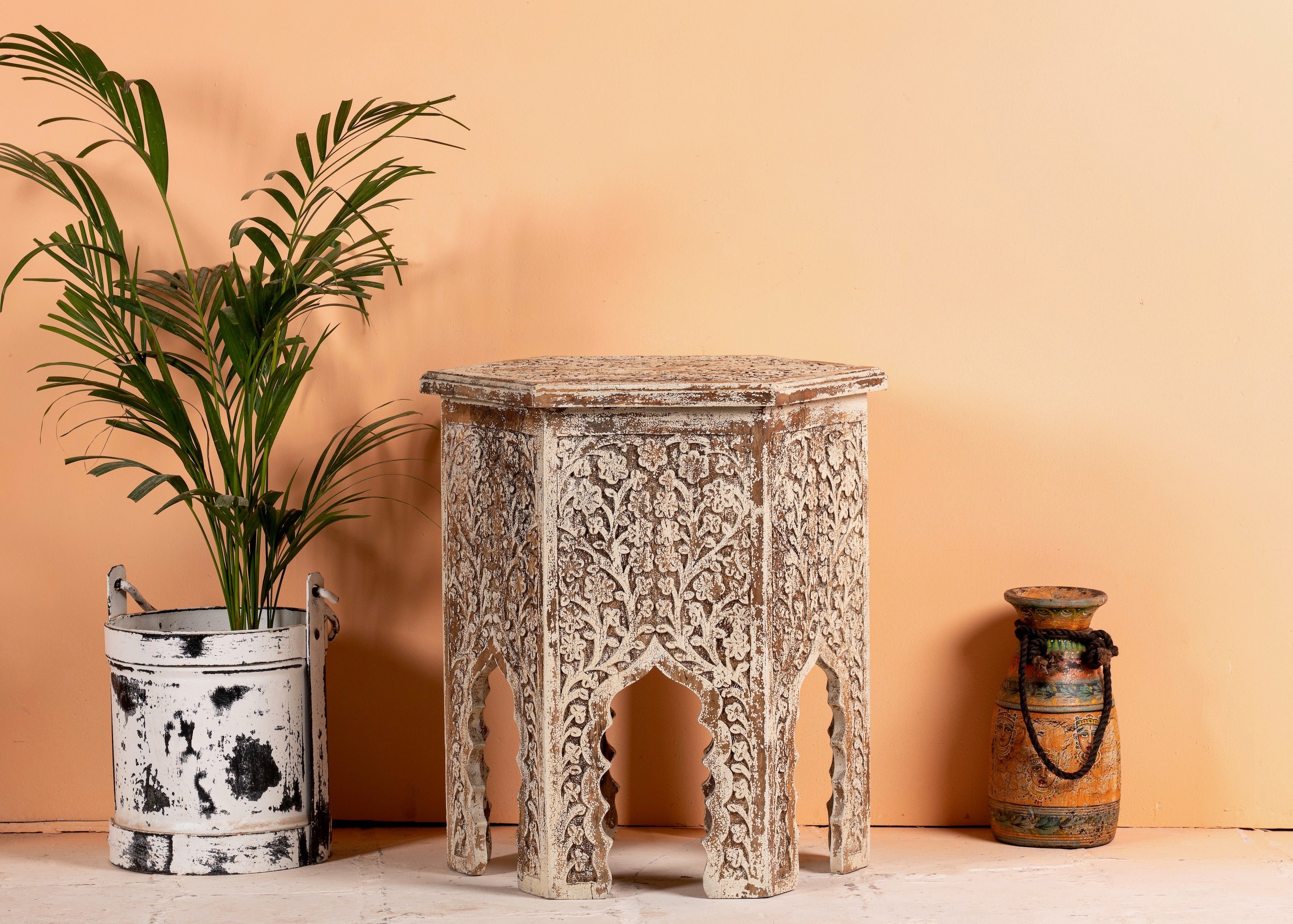 A hand-carved mango wood coffee table with a hexagonal shape and intricate floral and geometric carvings, displayed next to a potted plant and a vintage-style bucket.