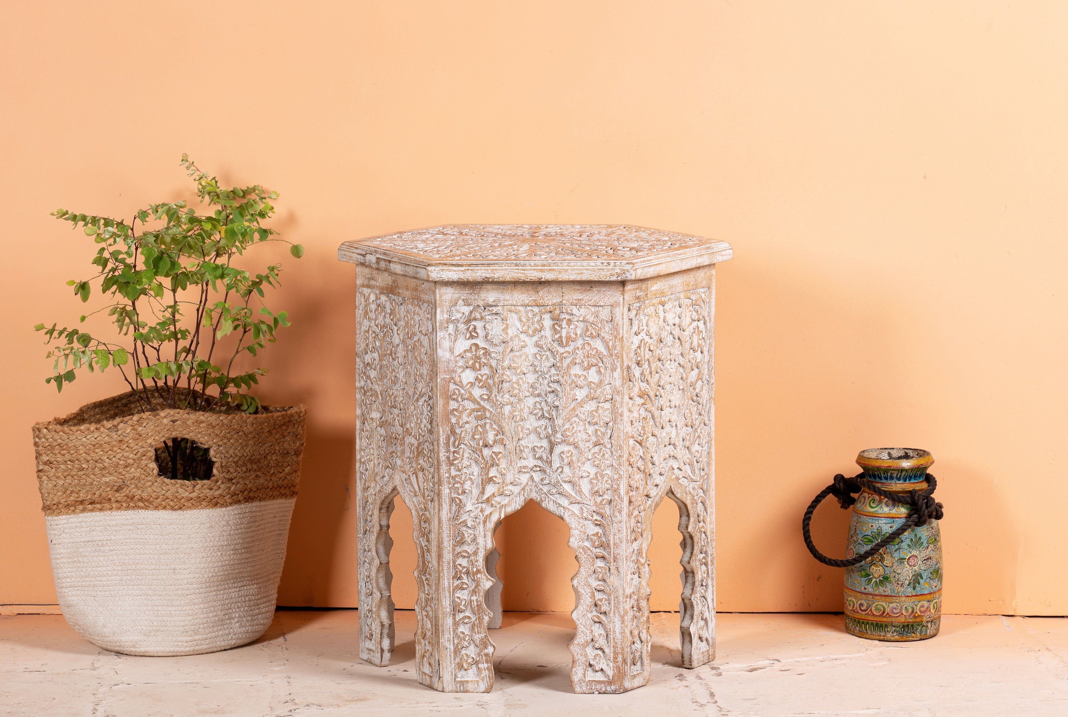A hand-carved, whitewashed hexagonal coffee table with Moroccan design details, placed against a neutral background with a plant to the left.