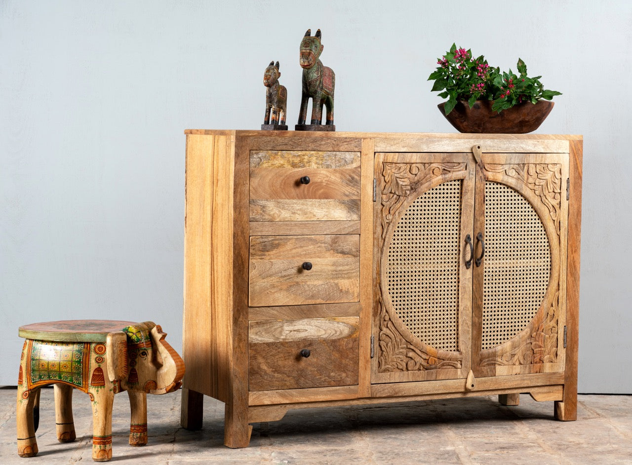 Side angle image of a hand carved mango wood cabinet in natural finish with 3 drawers and 2 rattan doors.