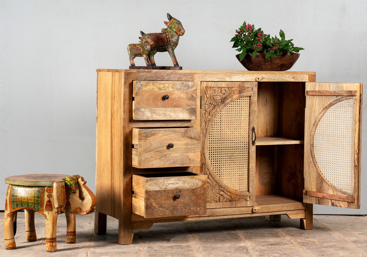 Side angle image with open doors of a hand carved mango wood cabinet in natural finish with 3 drawers and 2 rattan doors.