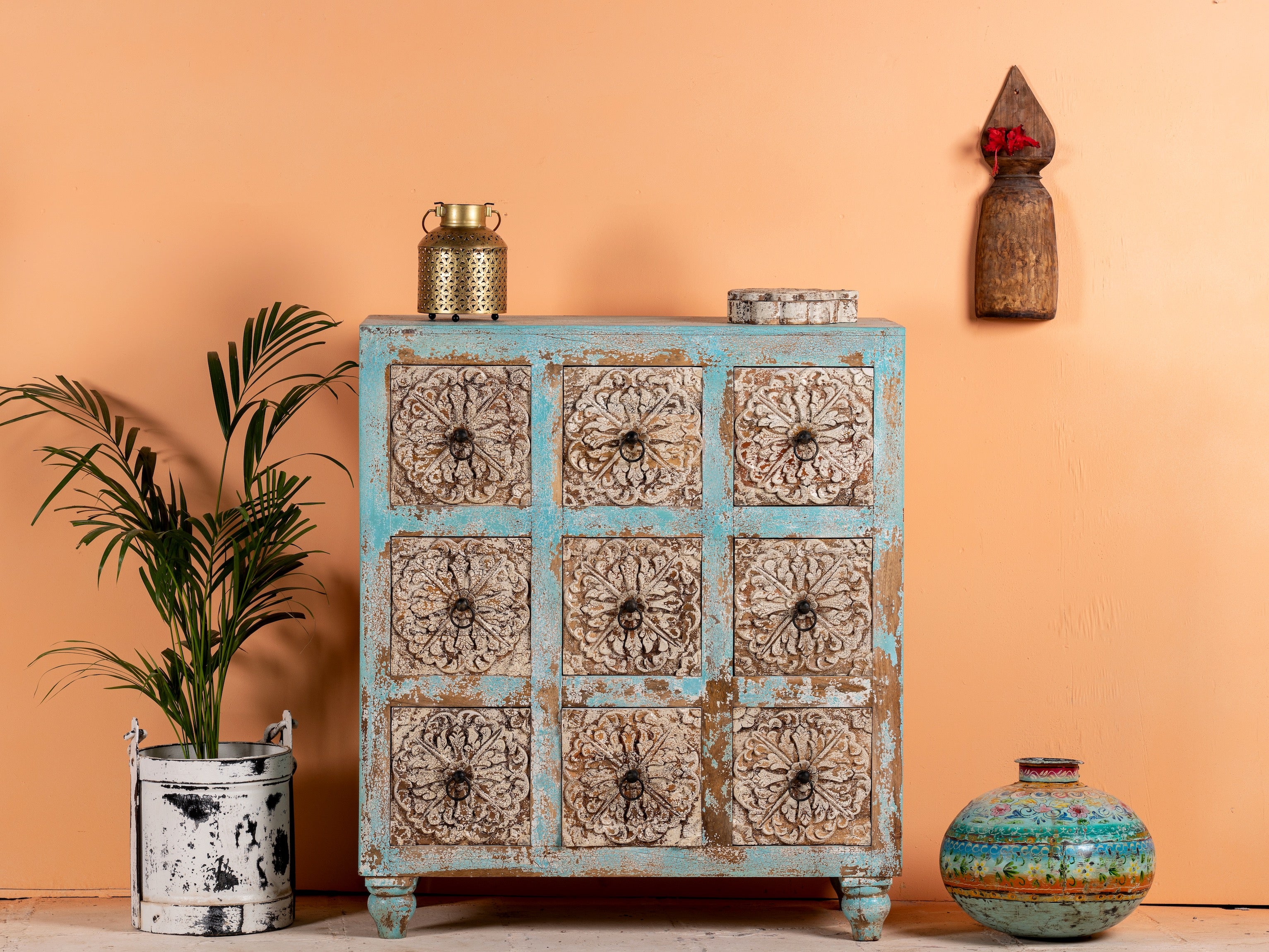 A vintage design mango wood chest of nine drawers with hand-carved floral patterns on the drawer fronts, featuring a blue body with whitewashed detailing and iron handles. The chest is positioned against a pink wall with a potted plant and decorative items on either side.