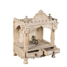 Side angle image of a decorative Indian altar made of mango wood, hand carved with 2 small drawers in a living room with a couch and coffee table.