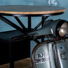 Close up image of a scooter bar table crafted from the front half of an original Bajaj scooter, featuring a round wooden top, metal frame, and rustic industrial finish.
