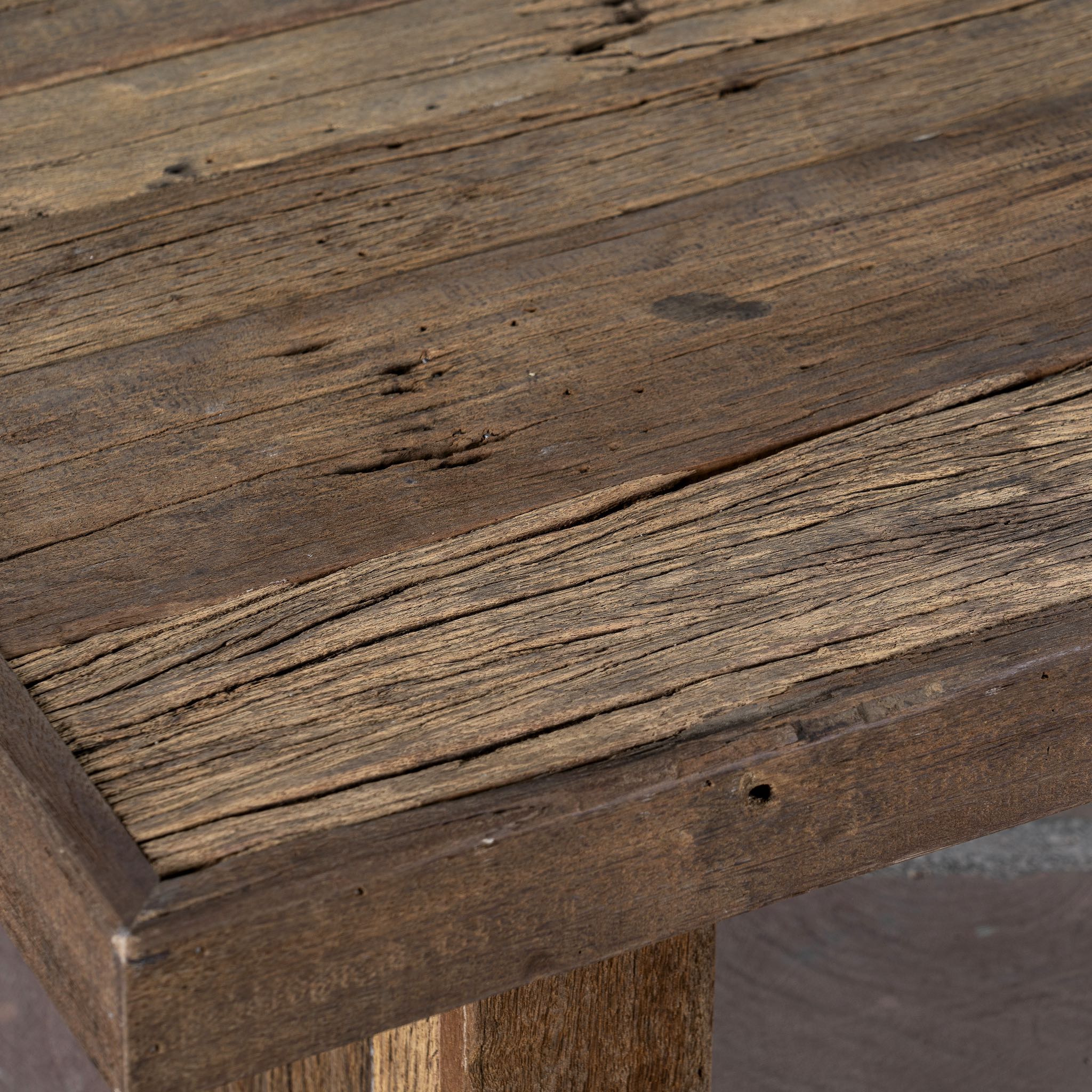 Close up image of a rectangular rustic coffee table constructed from reclaimed wood, featuring a thick plank-style top and wide block legs, all with a naturally weathered finish that highlights the wood’s grain and imperfections.

