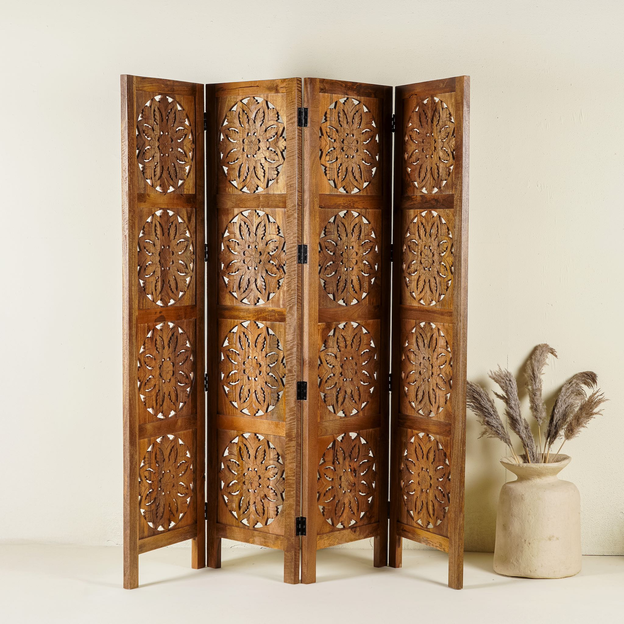 Foldable room partition made of mango wood, featuring four panels with symmetrical hand-carved circular floral medallions, finished in a rich natural wood tone and connected by black metal hinges for easy folding and adjustment.
