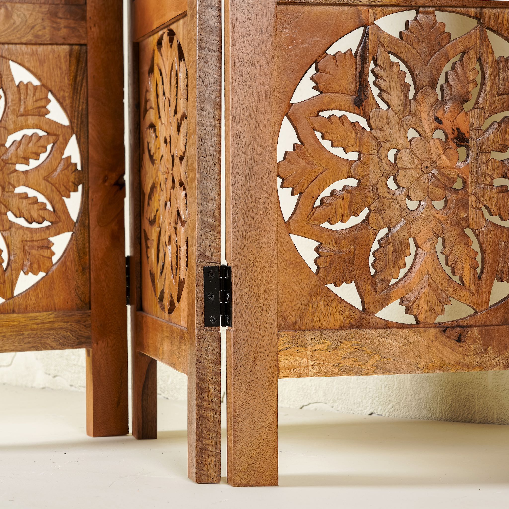 Close up image of a foldable room partition made of mango wood, featuring four panels with symmetrical hand-carved circular floral medallions, finished in a rich natural wood tone and connected by black metal hinges for easy folding and adjustment.
