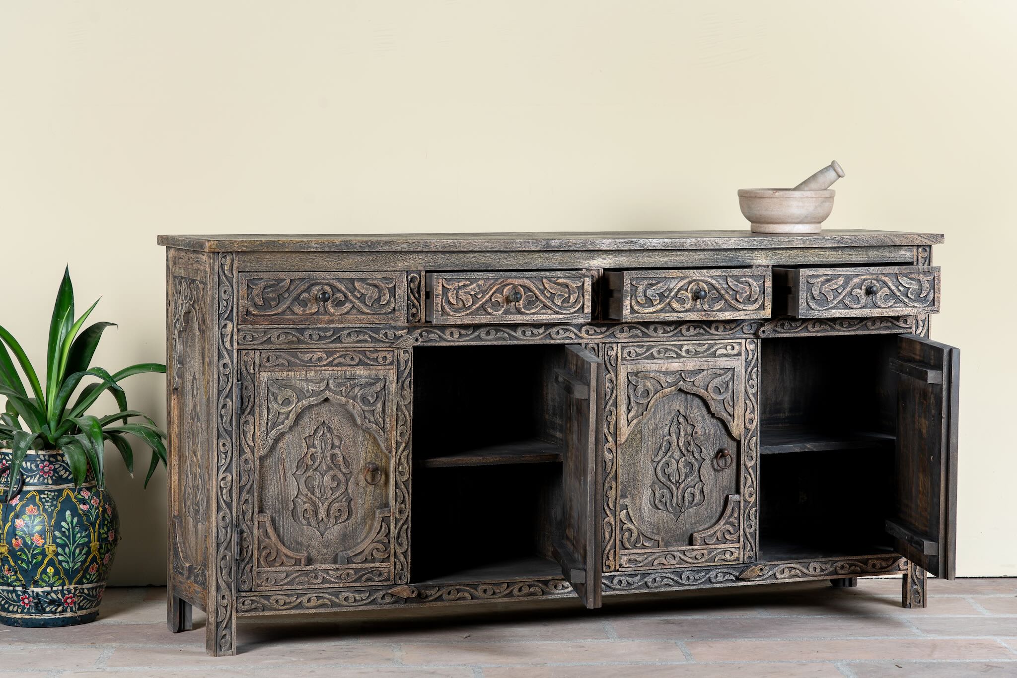 Side angle image with open doors of a hand-carved mango wood sideboard Aavya in a vintage dark finish, featuring four drawers and four carved cabinet doors with Indian-inspired motifs. A bold piece of vintage furniture and unique Indian craftsmanship.
