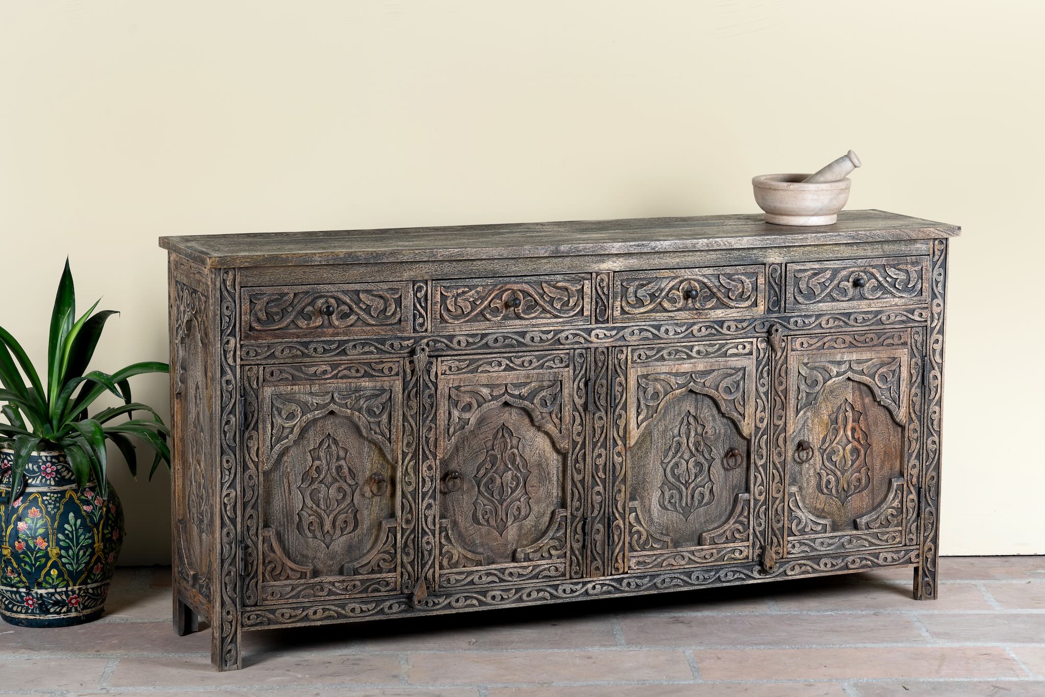 Side angle image of a hand-carved mango wood sideboard Aavya in a vintage dark finish, featuring four drawers and four carved cabinet doors with Indian-inspired motifs. A bold piece of vintage furniture and unique Indian craftsmanship.