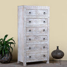 Side angle image of a hand carved white washed mango wood chest of 6 drawers in mediterranean design with iron 
handles.